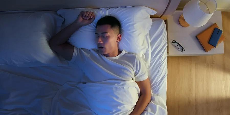 man sleeping on in a bed with his right hand up and his mouth open while wearing white shirt covered in white blanket