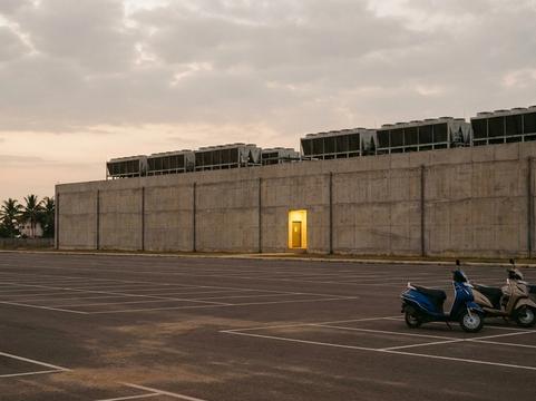 A nearly empty car park at dusk beside a windowless concrete data hall lined with rooftop cooling units; three scooters are parked in one corner and a single doorway is lit.
