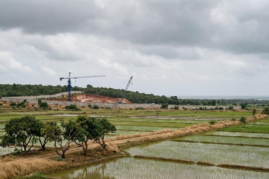 Flooded paddy fields and a small grove of trees in the foreground; behind them a long grey boundary wall, a cleared red-earth hillside and two construction cranes, one blue, with a grey sea on the horizon under monsoon cloud.
