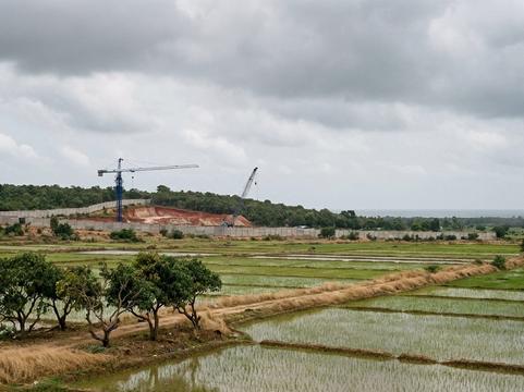 Flooded paddy fields and a small grove of trees in the foreground; behind them a long grey boundary wall, a cleared red-earth hillside and two construction cranes, one blue, with a grey sea on the horizon under monsoon cloud.