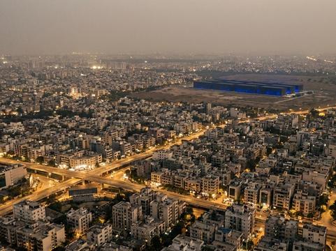 Aerial view of a dense Indian city at dusk with lit streets and a flyover, and at its far edge a long, low, windowless campus of halls glowing faintly blue on cleared land.