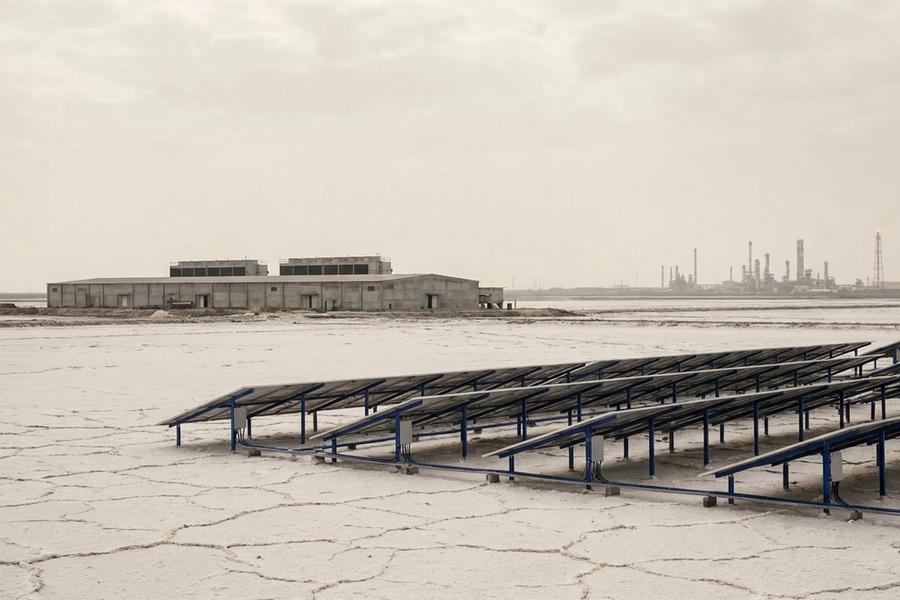 A cracked white salt flat with rows of solar panels on blue frames in the foreground, a long low concrete industrial hall in the middle distance, and refinery towers and a small flare on the horizon under an overcast sky.