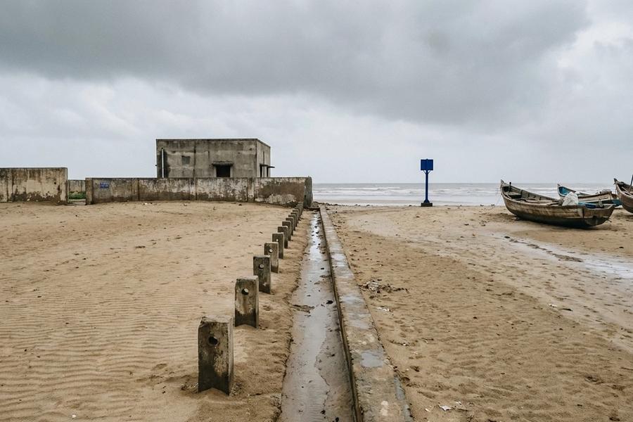 A sandy beach under a grey monsoon sky with a concrete channel and a row of short concrete markers running from the waterline to a small windowless concrete building behind a wall; wooden fishing boats rest on the sand and a blue marker post stands at the water's edge.