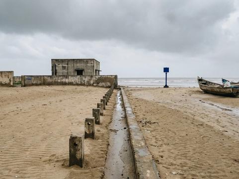 A sandy beach under a grey monsoon sky with a concrete channel and a row of short concrete markers running from the waterline to a small windowless concrete building behind a wall; wooden fishing boats rest on the sand and a blue marker post stands at the water's edge.
