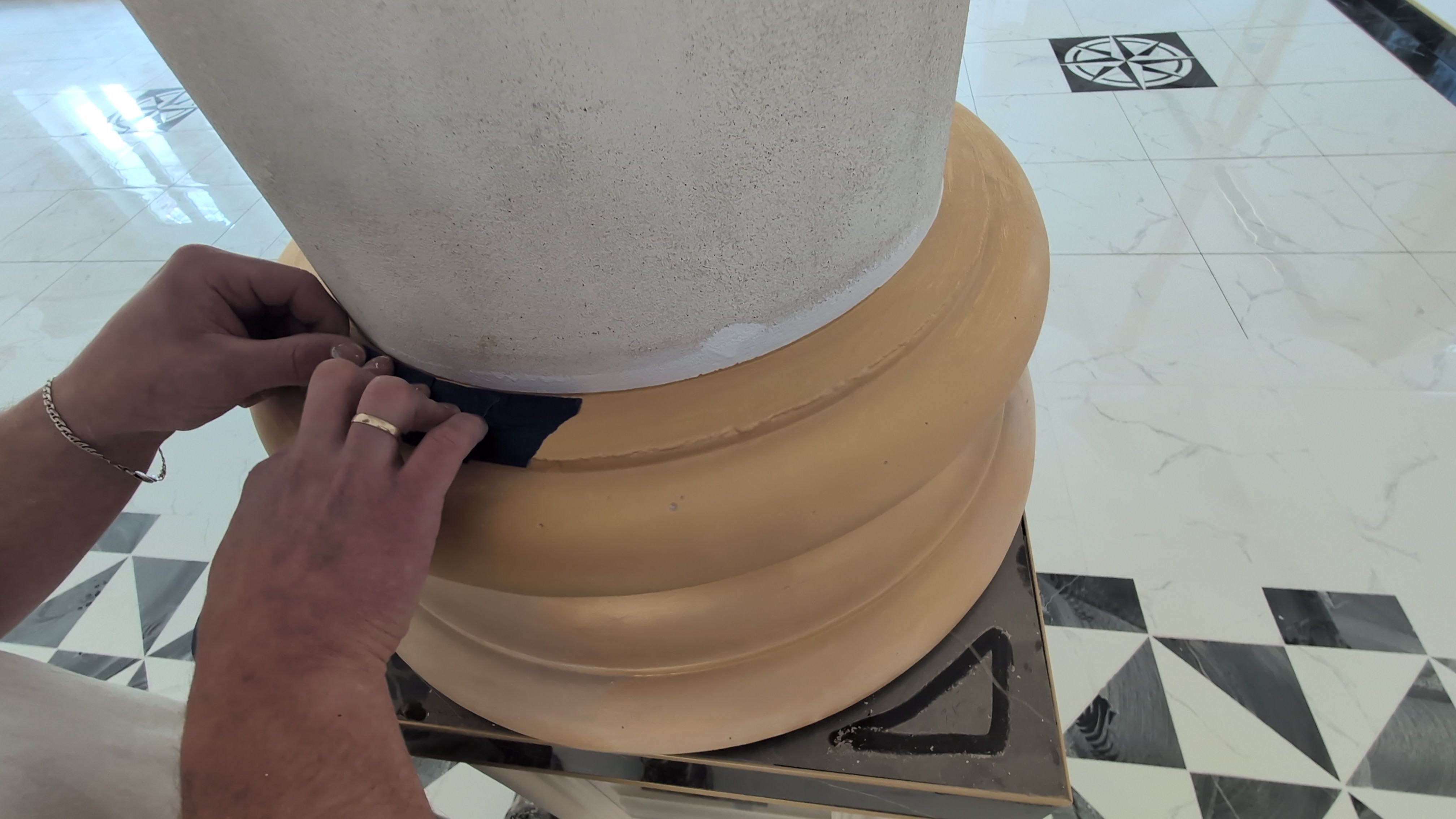 Mężczyzna zabezpieczający taśmą malarską  bazę kolumny przed pokrywaniem gładzią.. Na zdjęciu widoczne dłonie przyklejające  niebieską taśmę do bazy kolumny, w tle flizy na posadzce składająca się z biało-czarnych trójkątów 