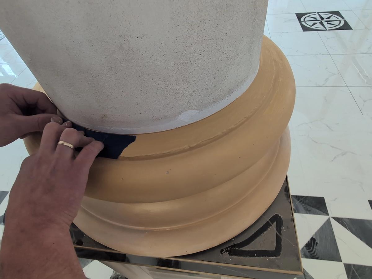 Mężczyzna zabezpieczający taśmą malarską bazę kolumny przed pokrywaniem gładzią.. Na zdjęciu widoczne dłonie przyklejające niebieską taśmę do bazy kolumny, w tle flizy na posadzce składająca się z biało-czarnych trójkątów