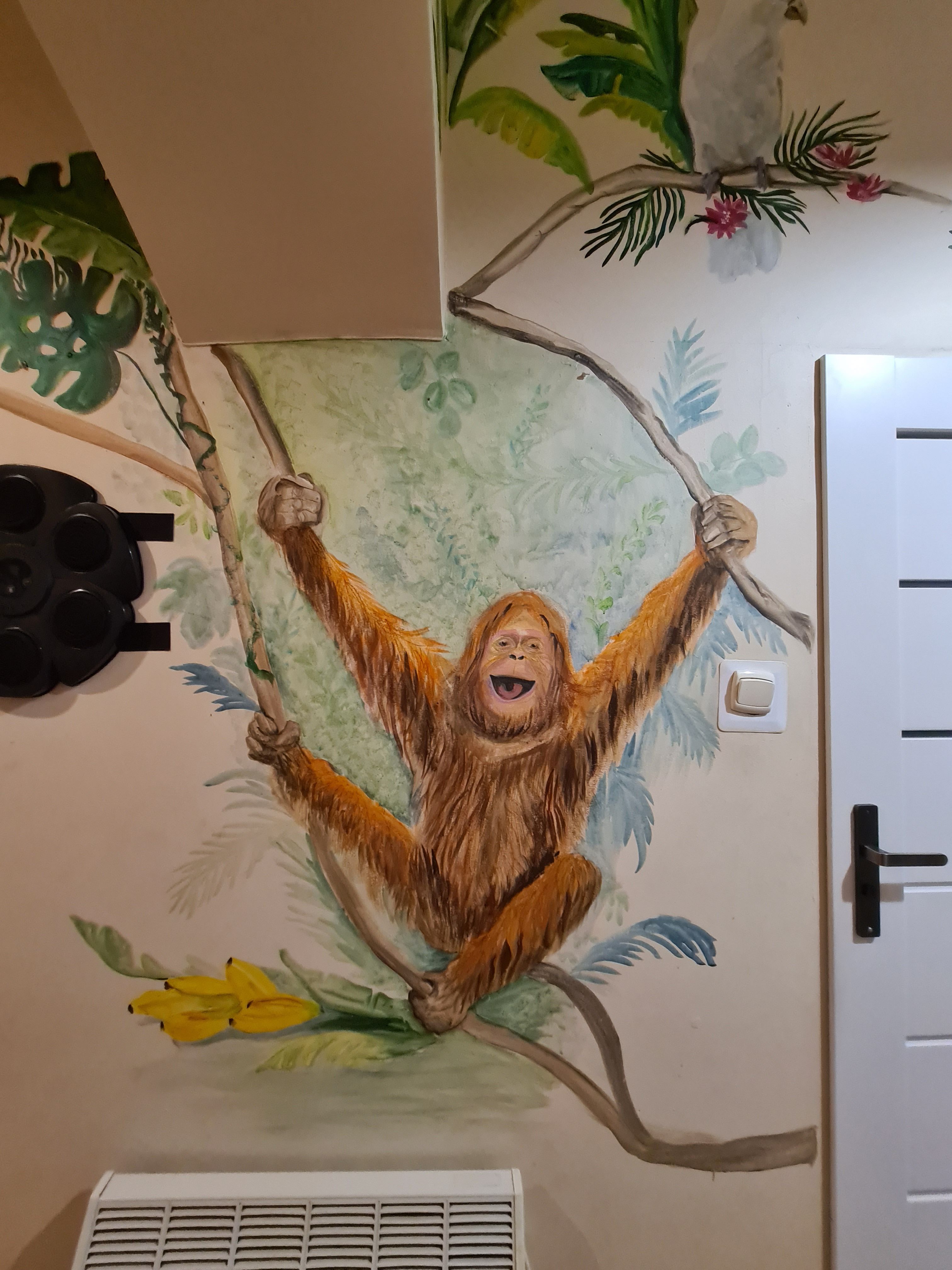 Uśmiechnięty orangutan hustający się wśród drzew. Dookoła tropikalna roślinność. Mural autorski.
