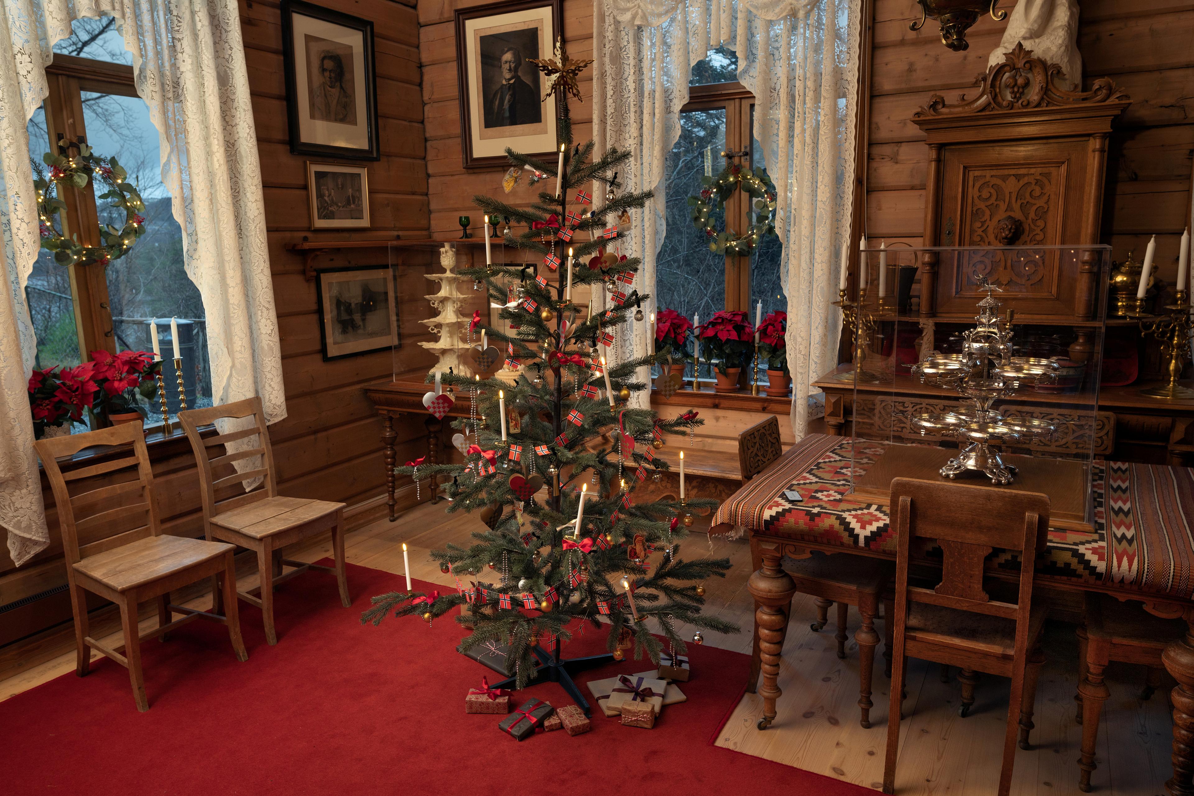 Et interiør fra Griegs villa som viser et juletre, pyntet med lys og norske flagg stående i spisestuen. I vinduene står juleblomster og julekranser er hengt opp. 