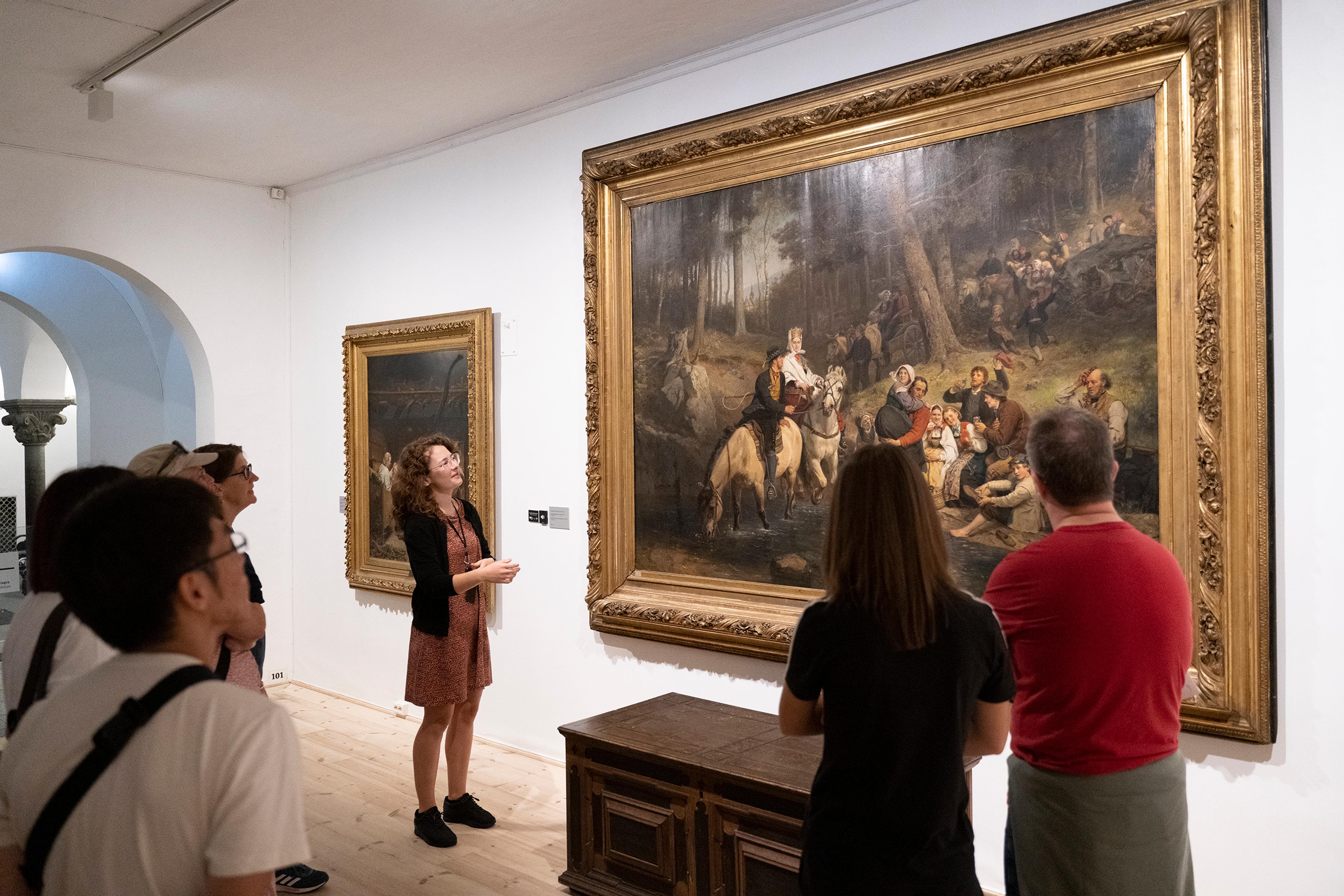 A museum guide is looking at a large painting, while talking to a group of visitors.