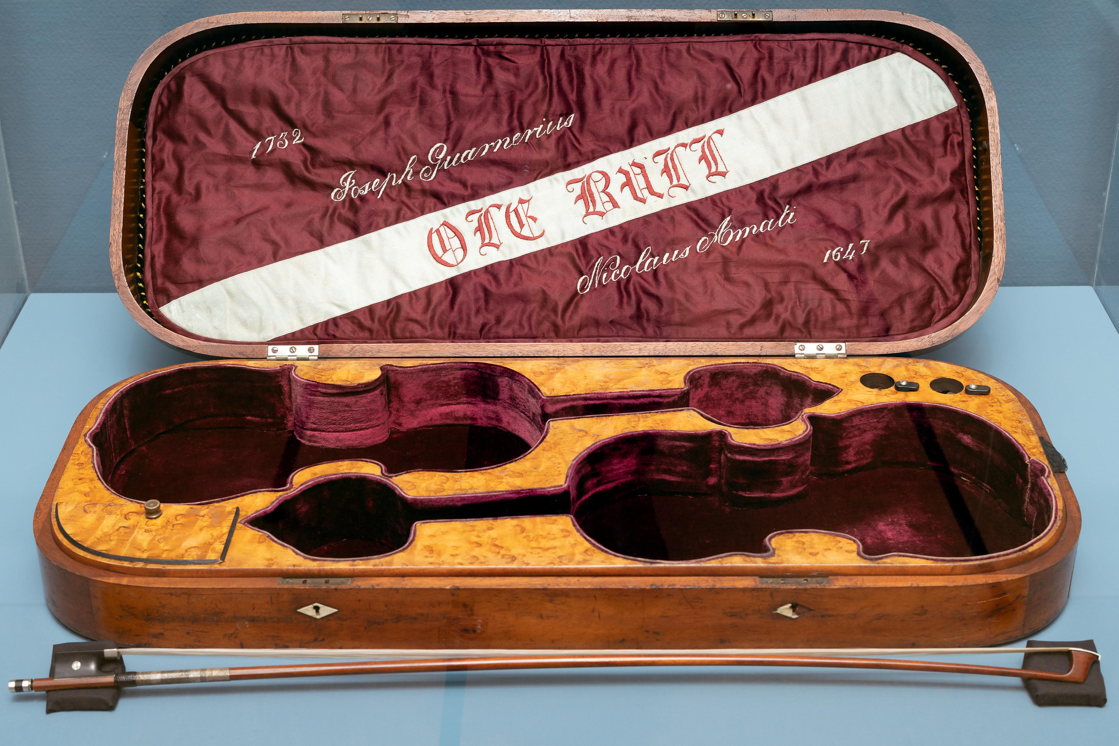 En brun fiolinkasse med rød tekstil på innsiden, og med Ole Bulls navn som motiv i lokket. Foran kassen ligger en bue.
