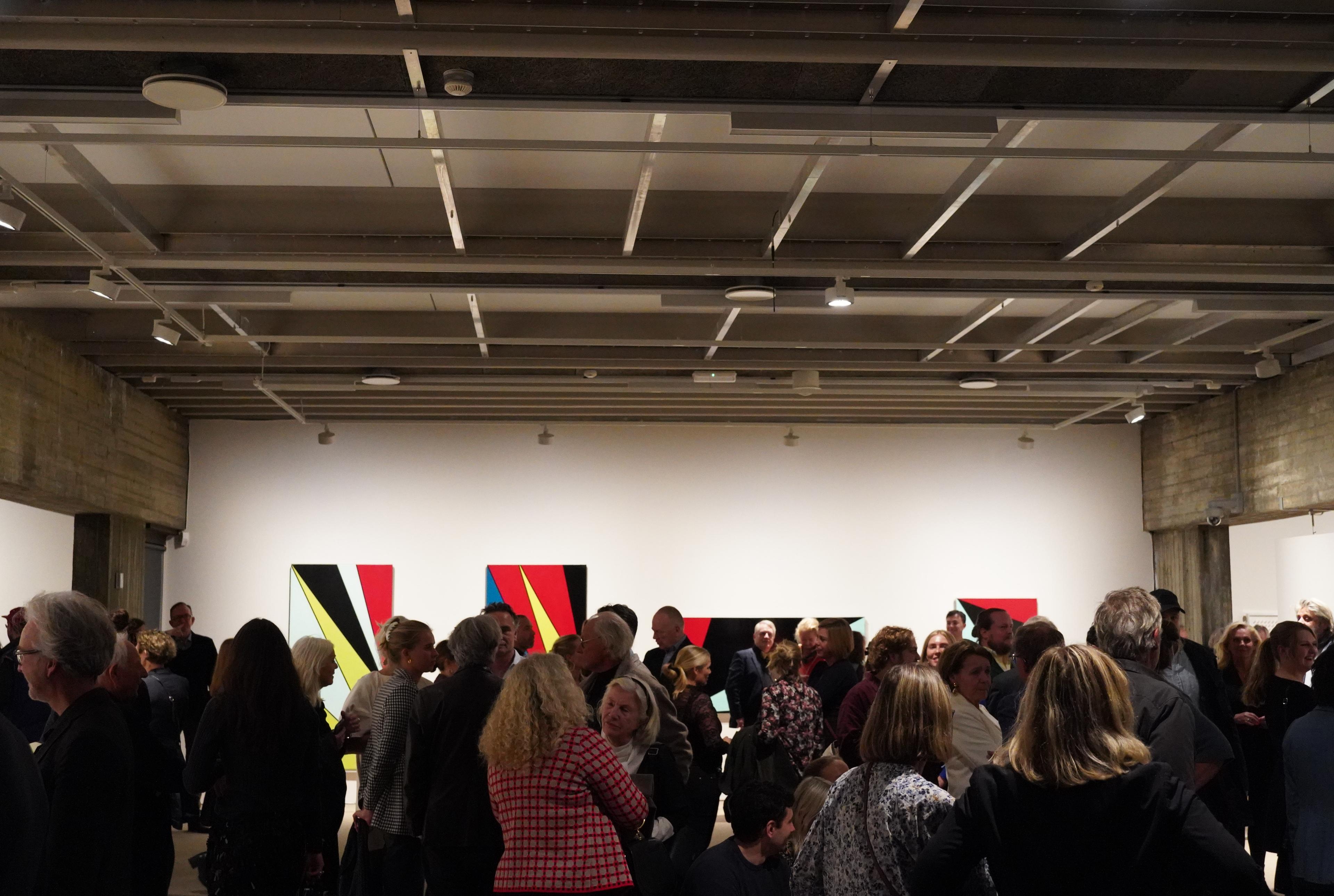 En utstillingssal full av folk, foran hvite vegger med abstrakte malerier