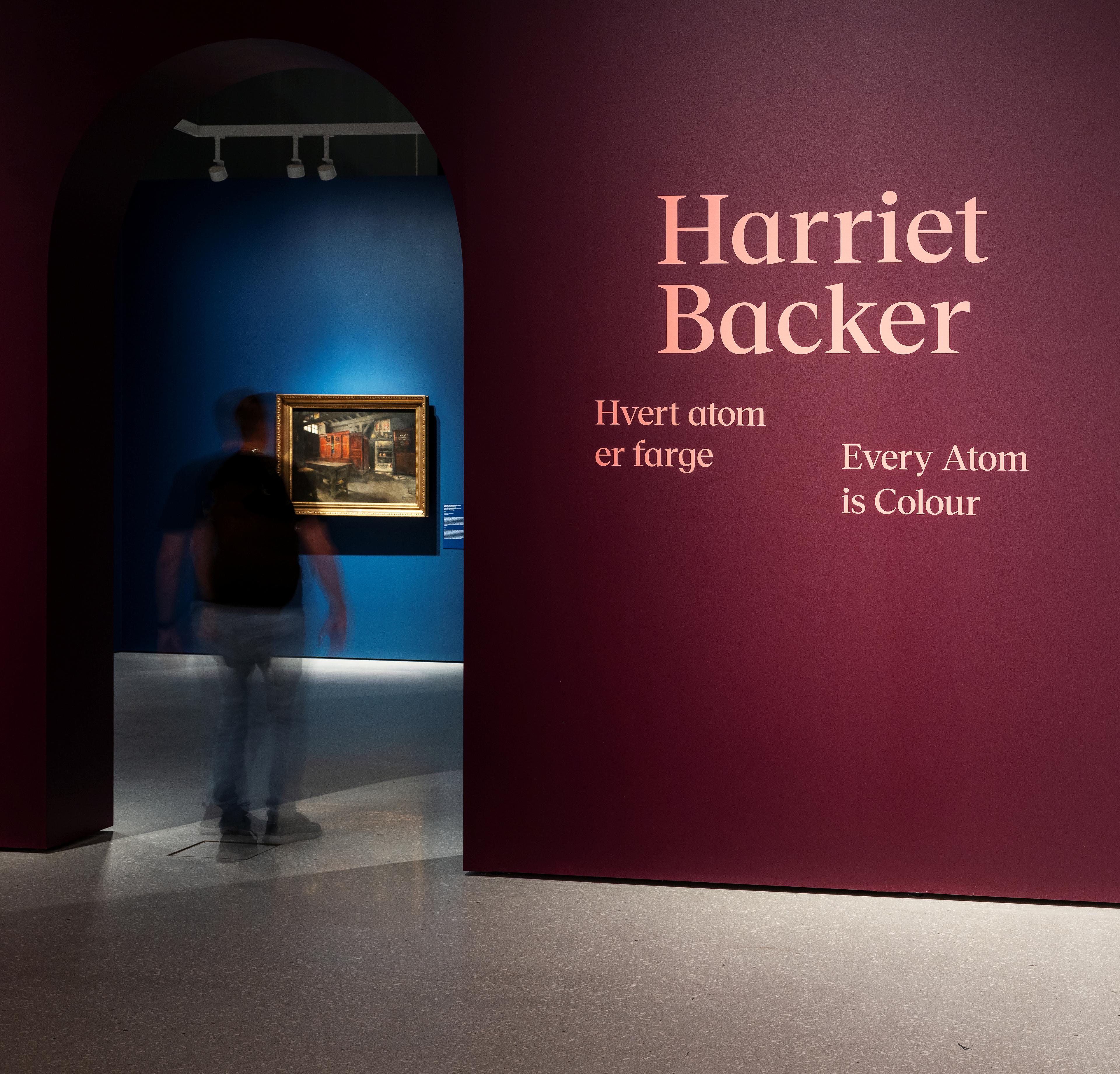 Et utstillingsrom med burgunder vegger og teksten Harriet Backer Hvert atom er farge