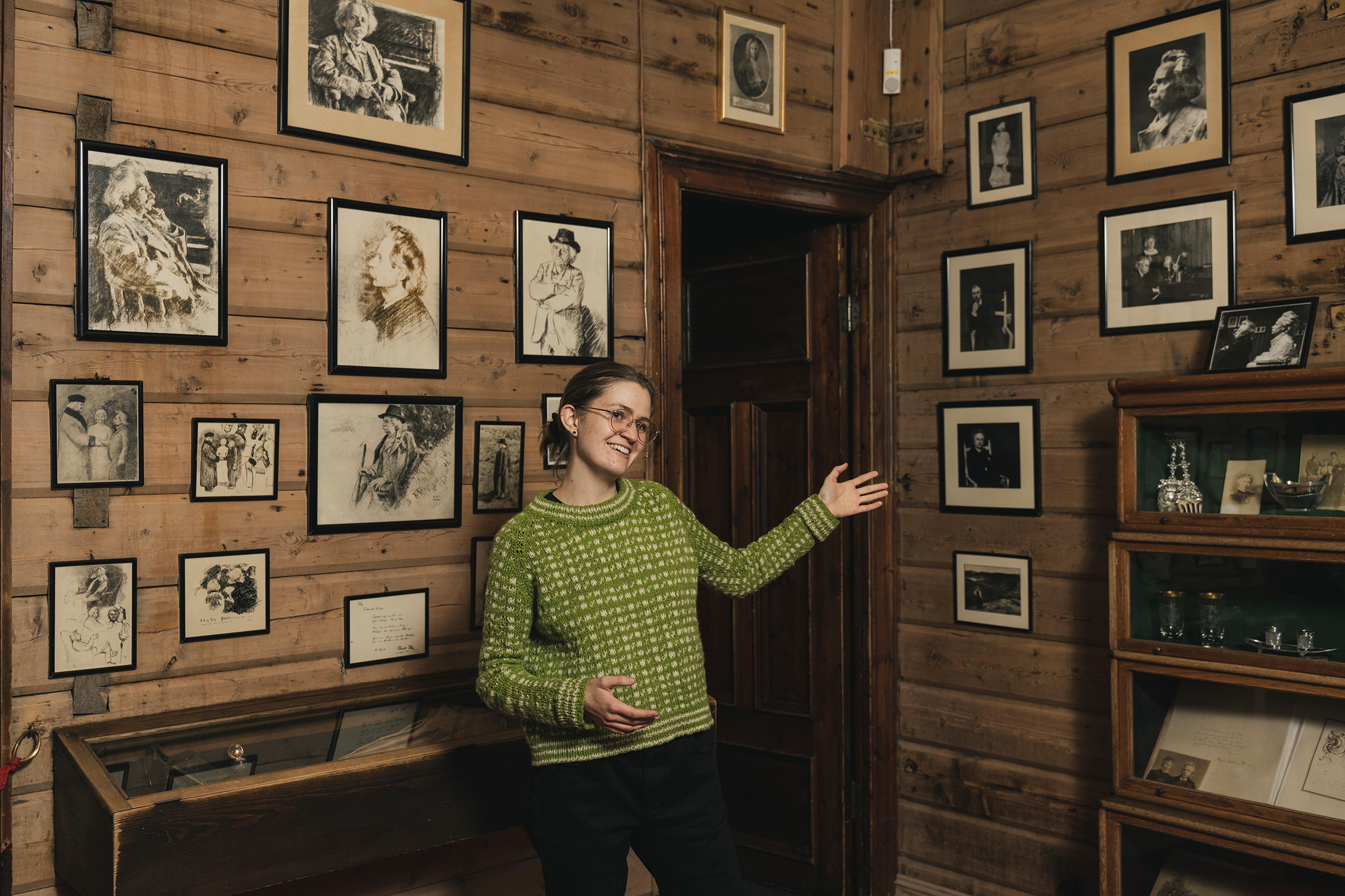 En omviser i Griegs villa på Troldhaugen peker mot en vegg med fotografier. Hun smiler mot kamera.