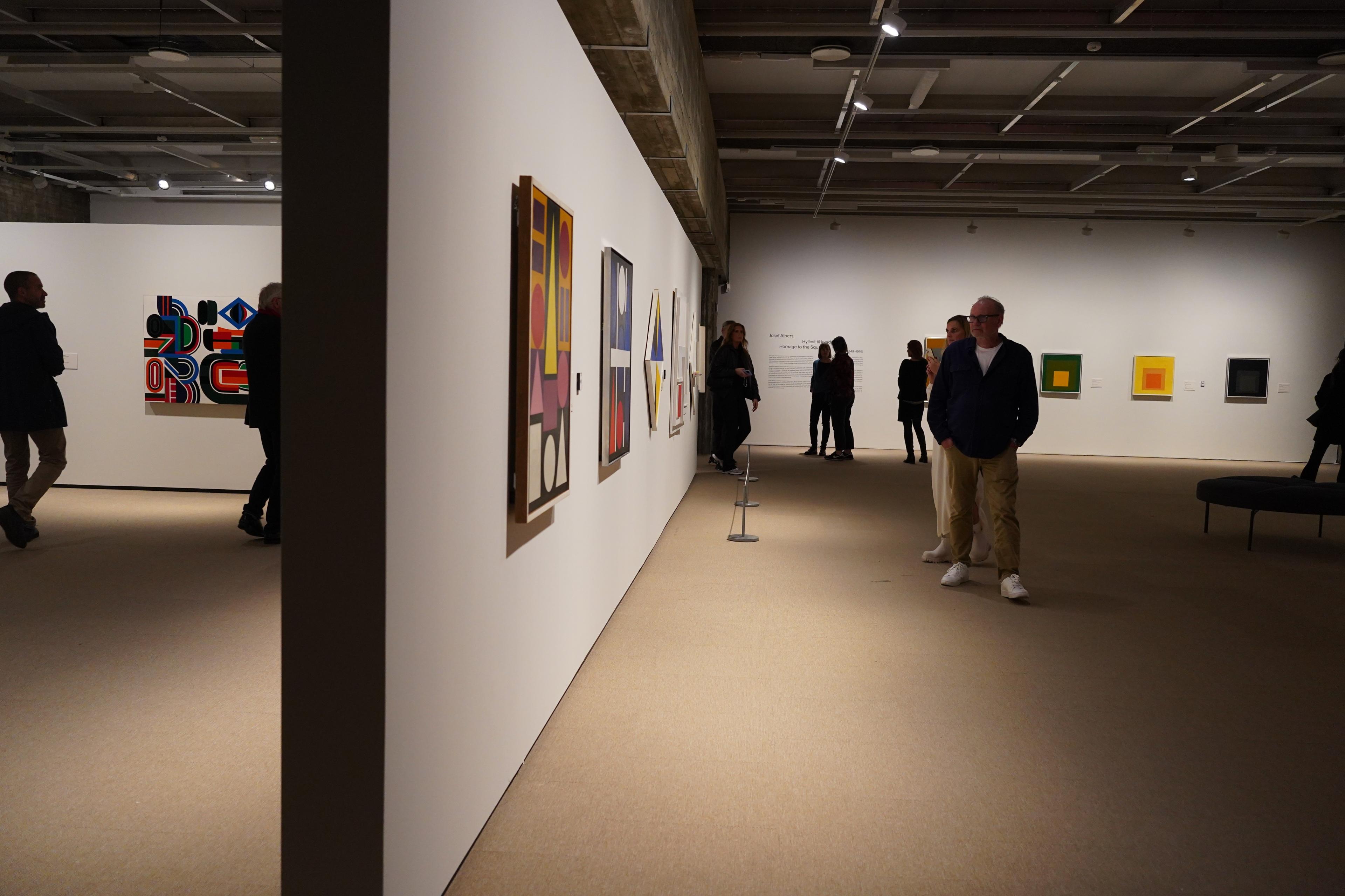 Personer i et utstillingsrom, med abstrakte malerier, som henger på en hvit utstillingsvegg