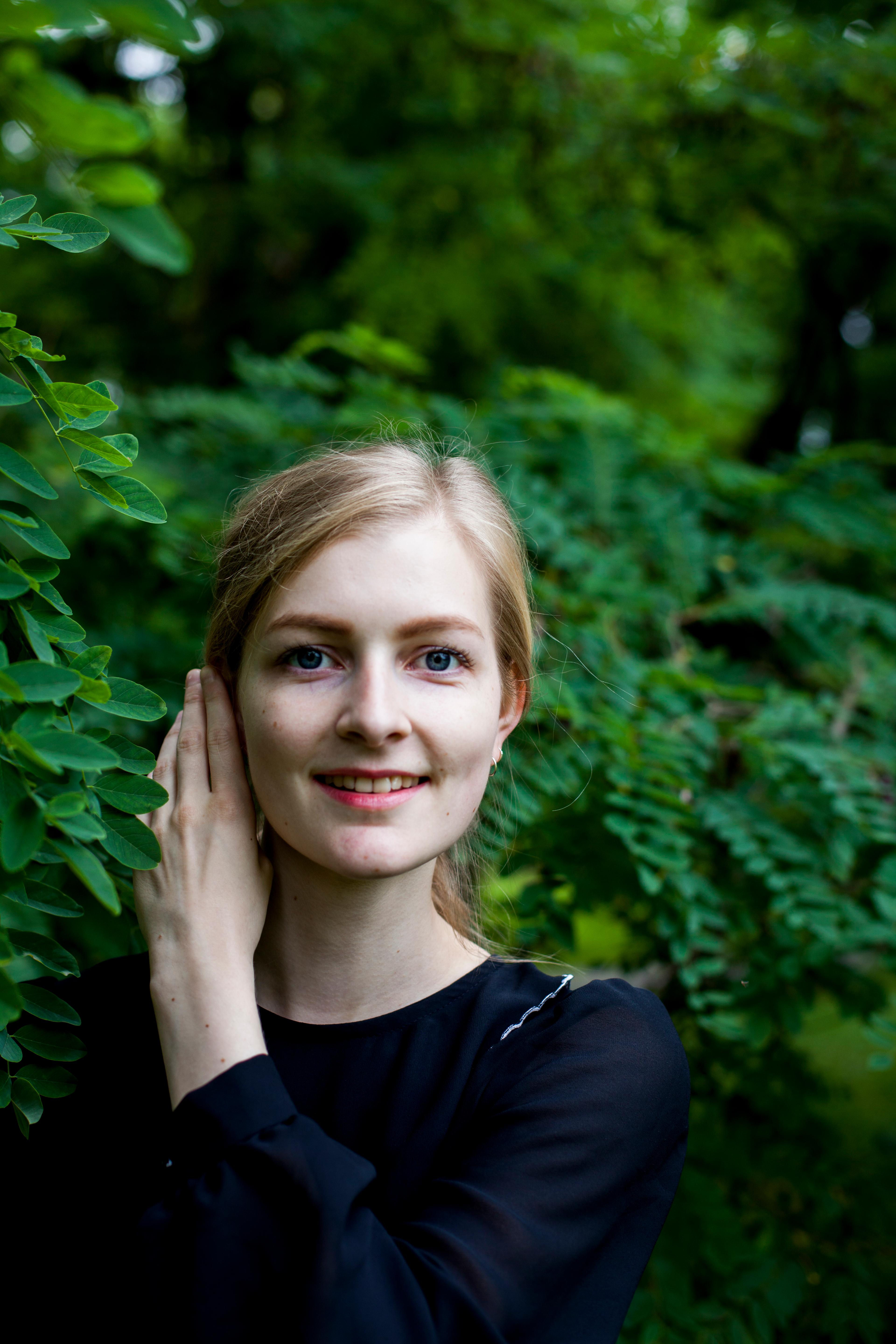 Portrett av pianisten Siril Valberg som står i en grønn skog. Hun ser rett frem og smiler, trekker håret bak øret med venstre hånd.