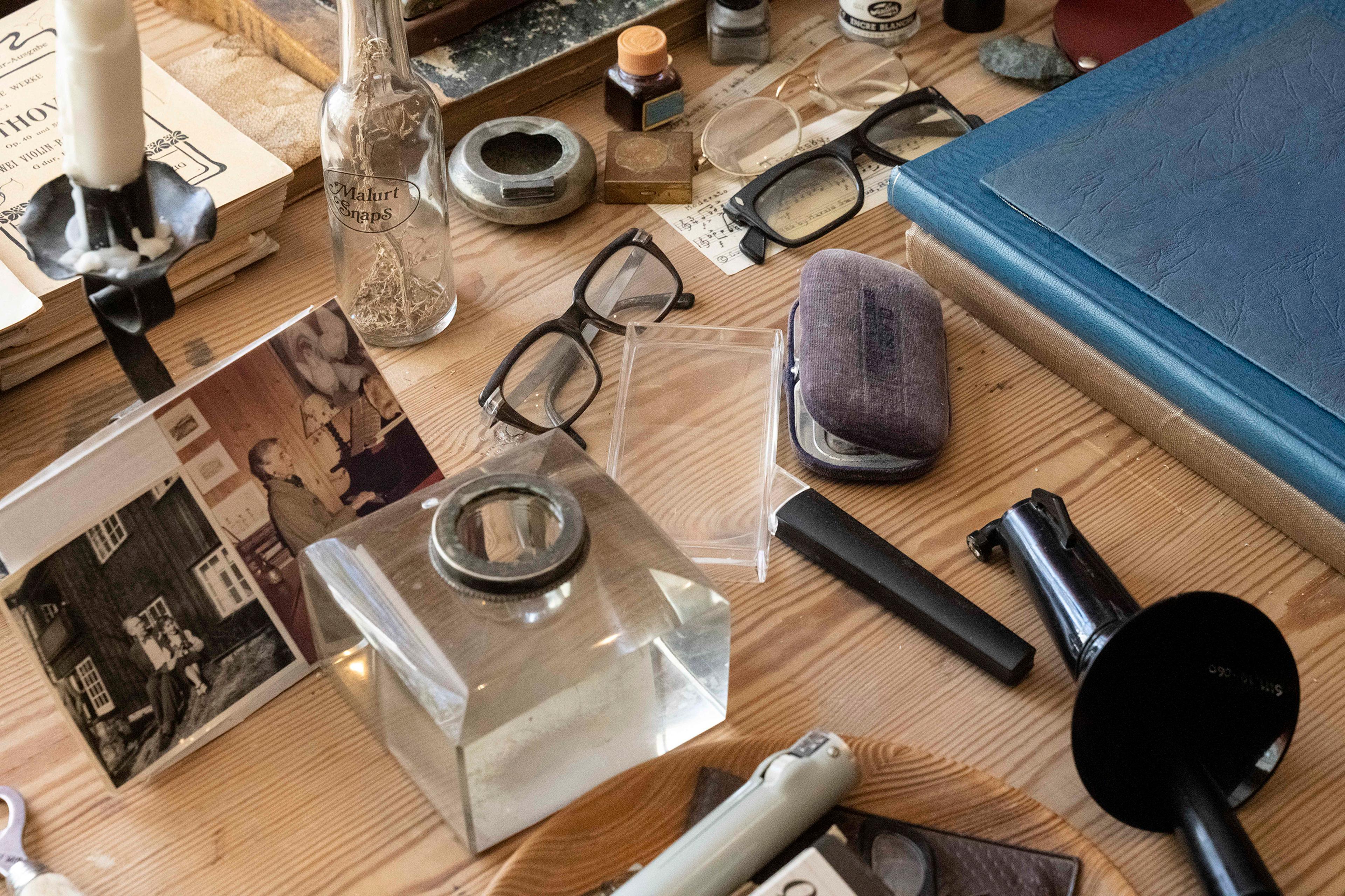Detaljer fra arbeidsbordet til Harald Sæverud. Vi ser blant annet en liten flaske med en urt oppi, flere brillepar og fotografier.