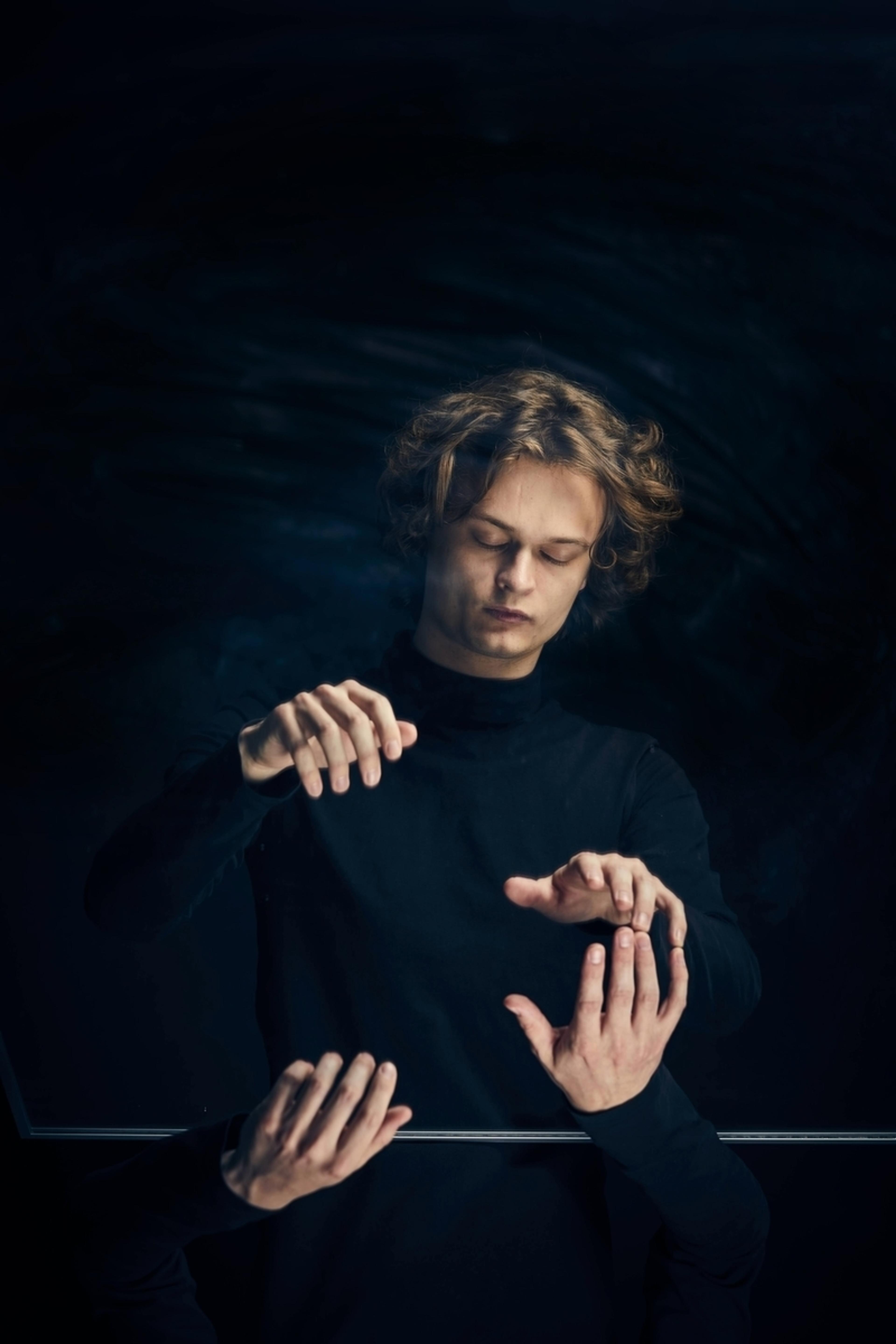 En ung mann gestikulerer med hendene, som han spiller på piano. Han er kledd i sort, mot en sort bakgrunn.
