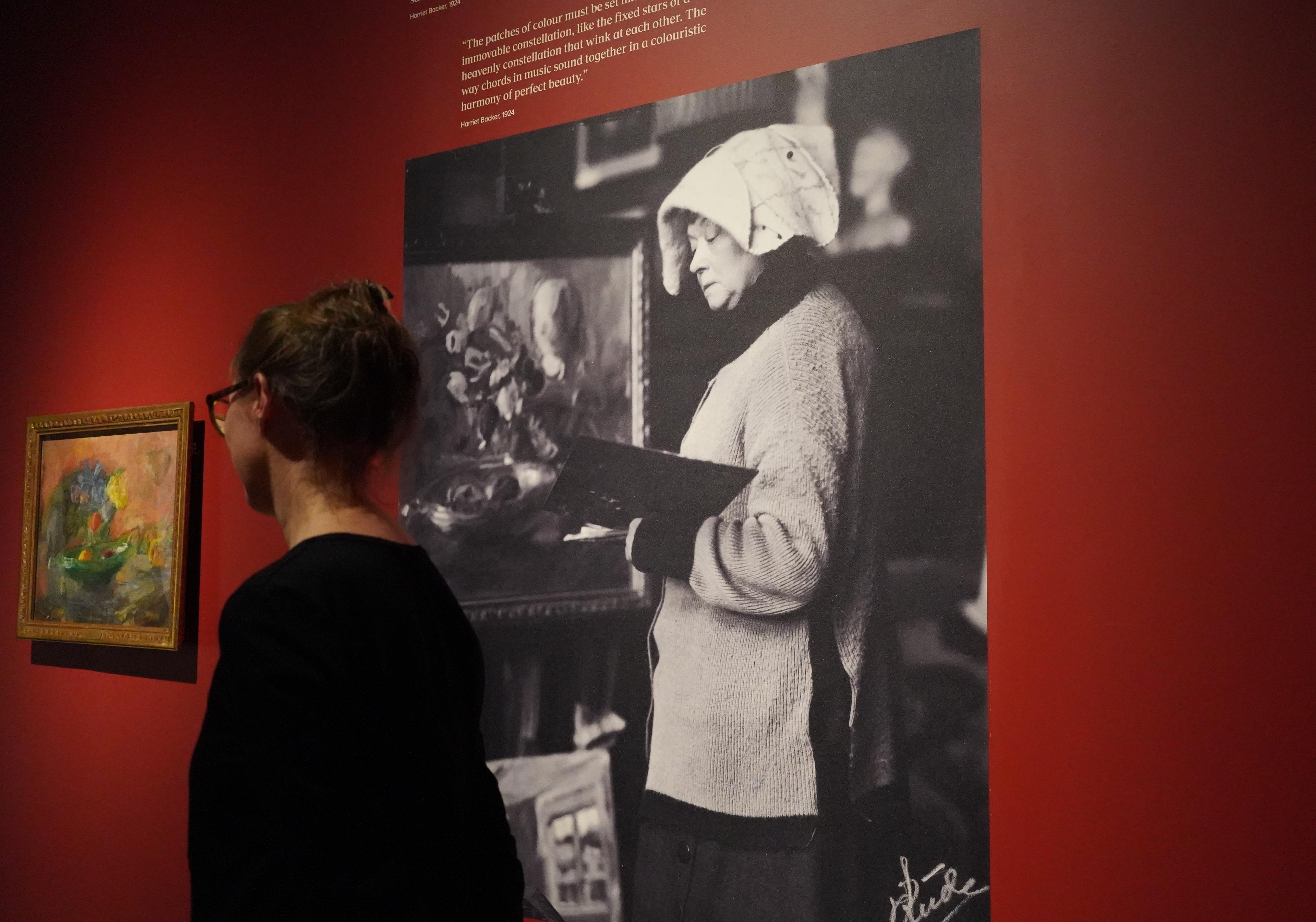 En kvinne står foran en rød utstillingsvegg, med et bilde av Harriet Backer på