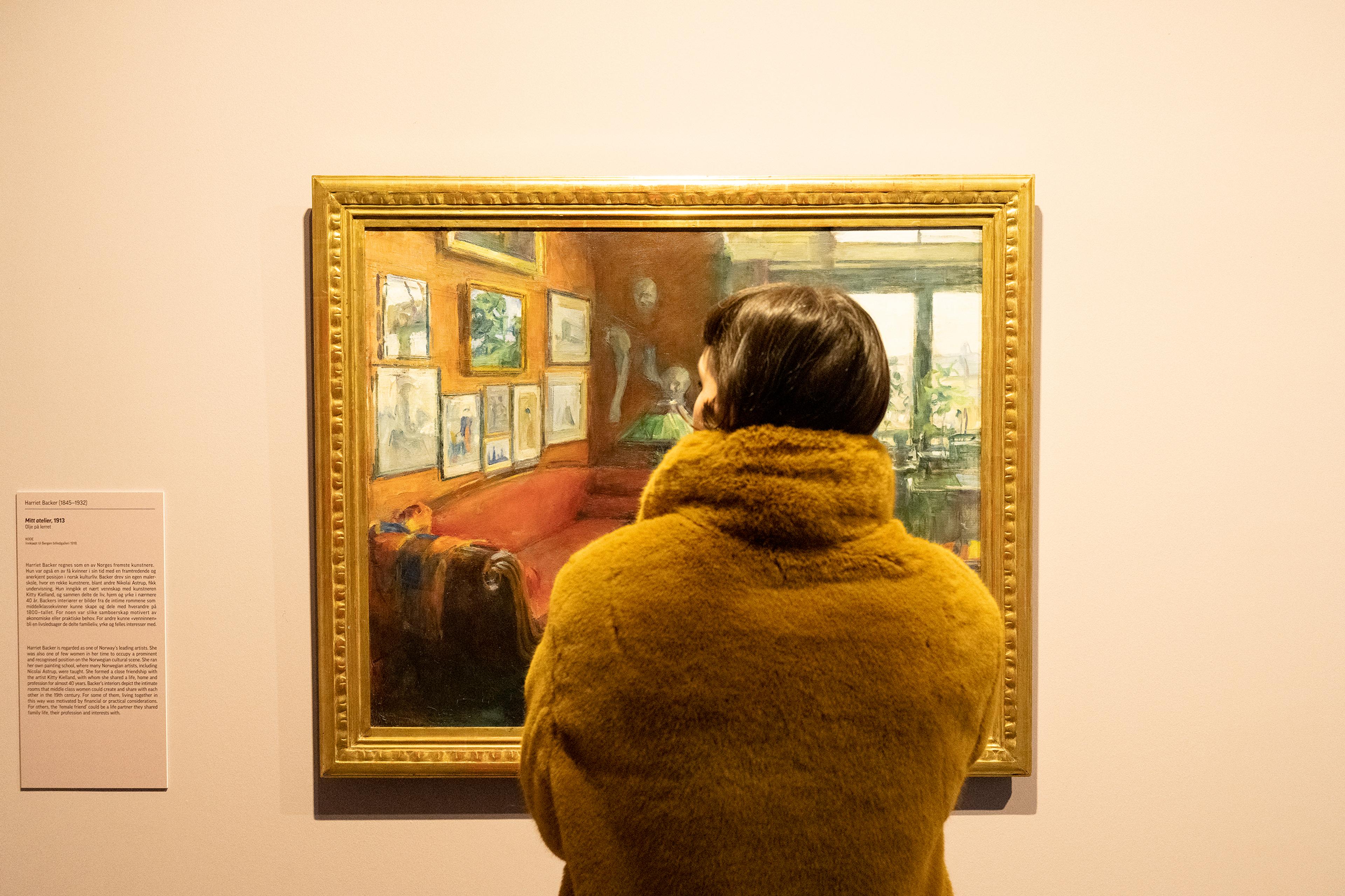 En person står foran et maleri av Harriet Backer, som henger på en vegg i en dus gul farge