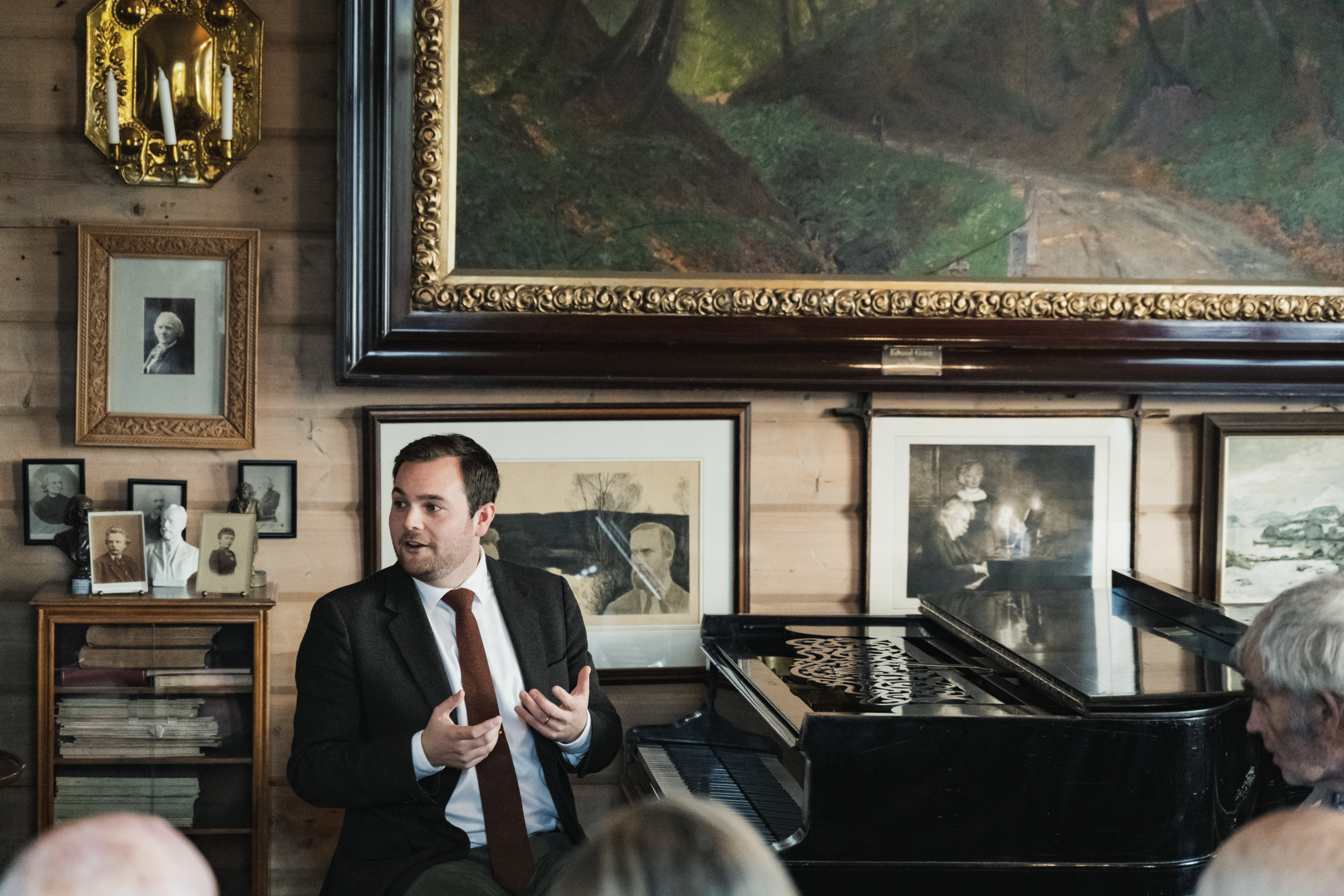 Foto som viser direktør ved komponisthjemmene, Christian Grøvlen, sittende ved pianoet i Griegs villa. Han forteller noe til publikum. 