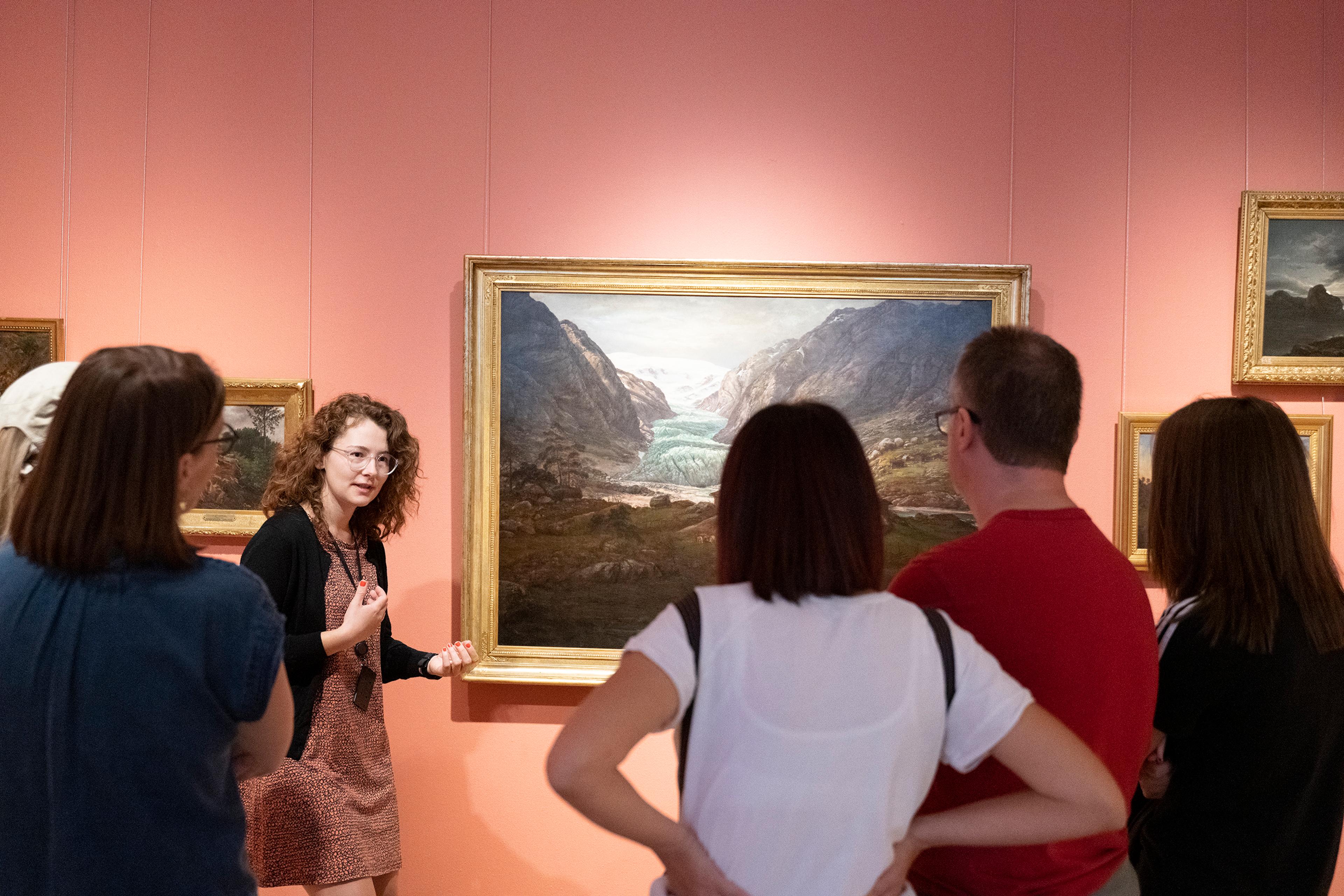 Et bilde som viser en gruppe menneskre på omvisning som ser på et maleri. 