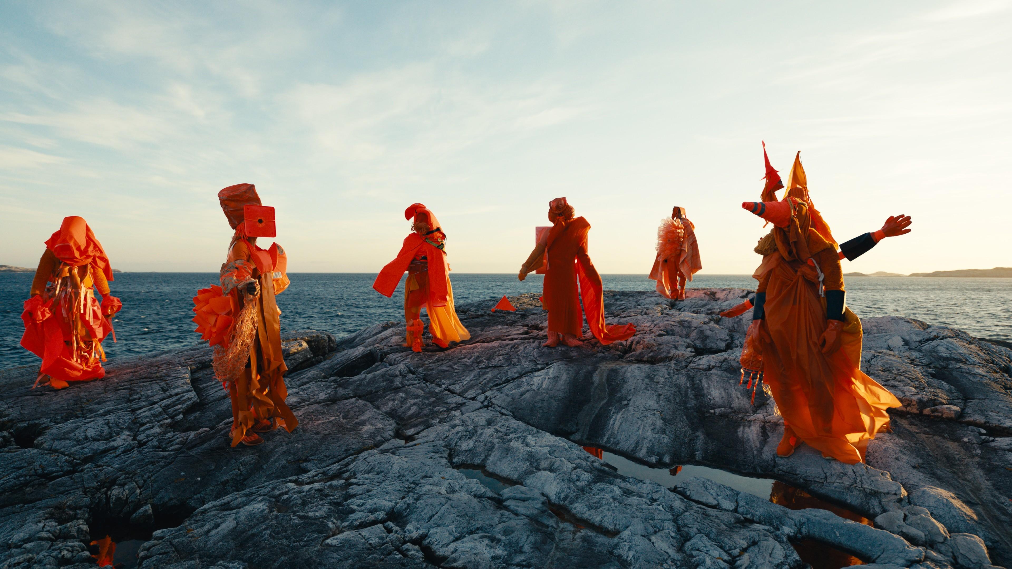 En videostill av dansende mennesker i oransje bølgende klær på en klippe