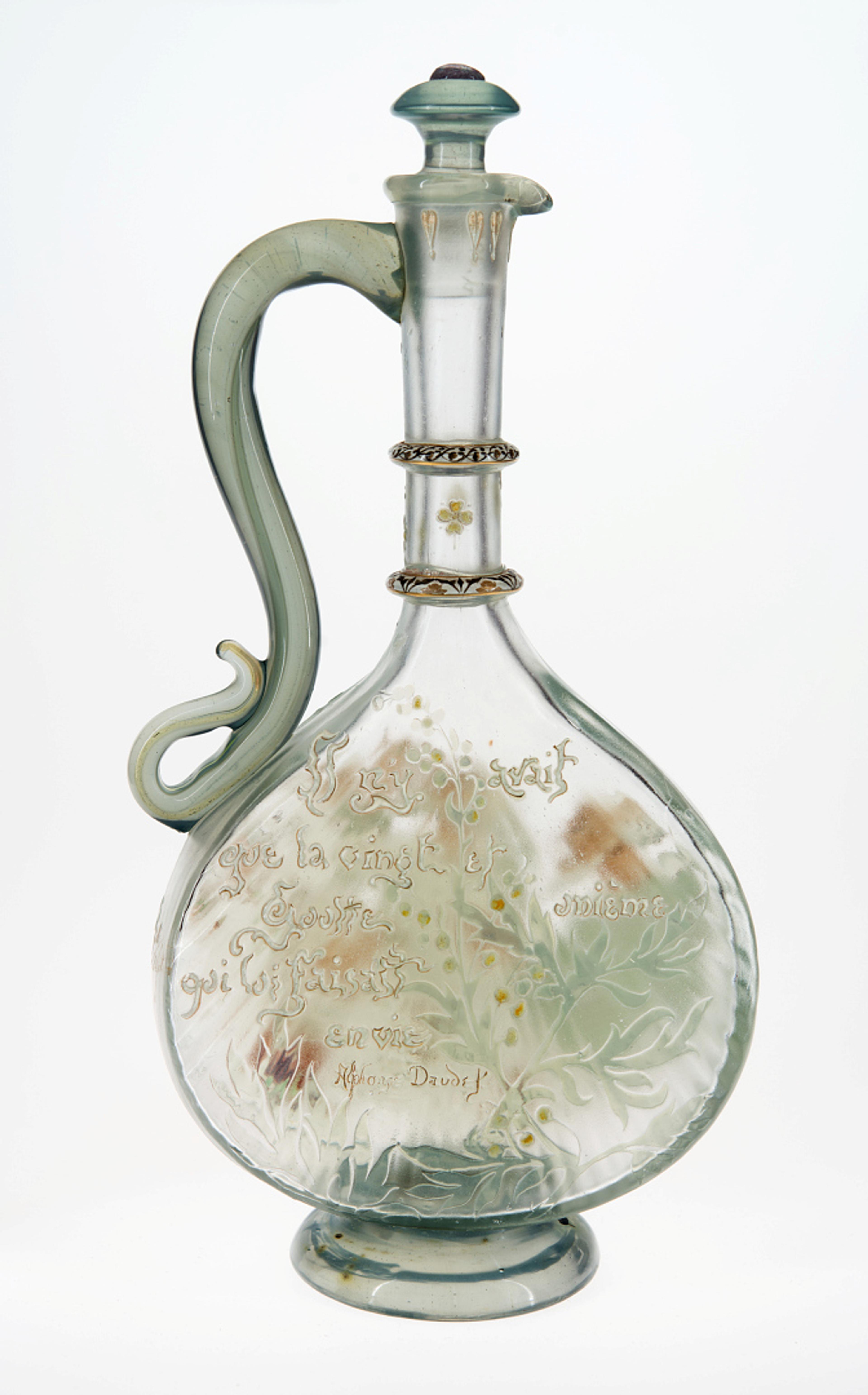 Foto av en vase i glass, med en smal hals og buet hank, med grønnskjær.
