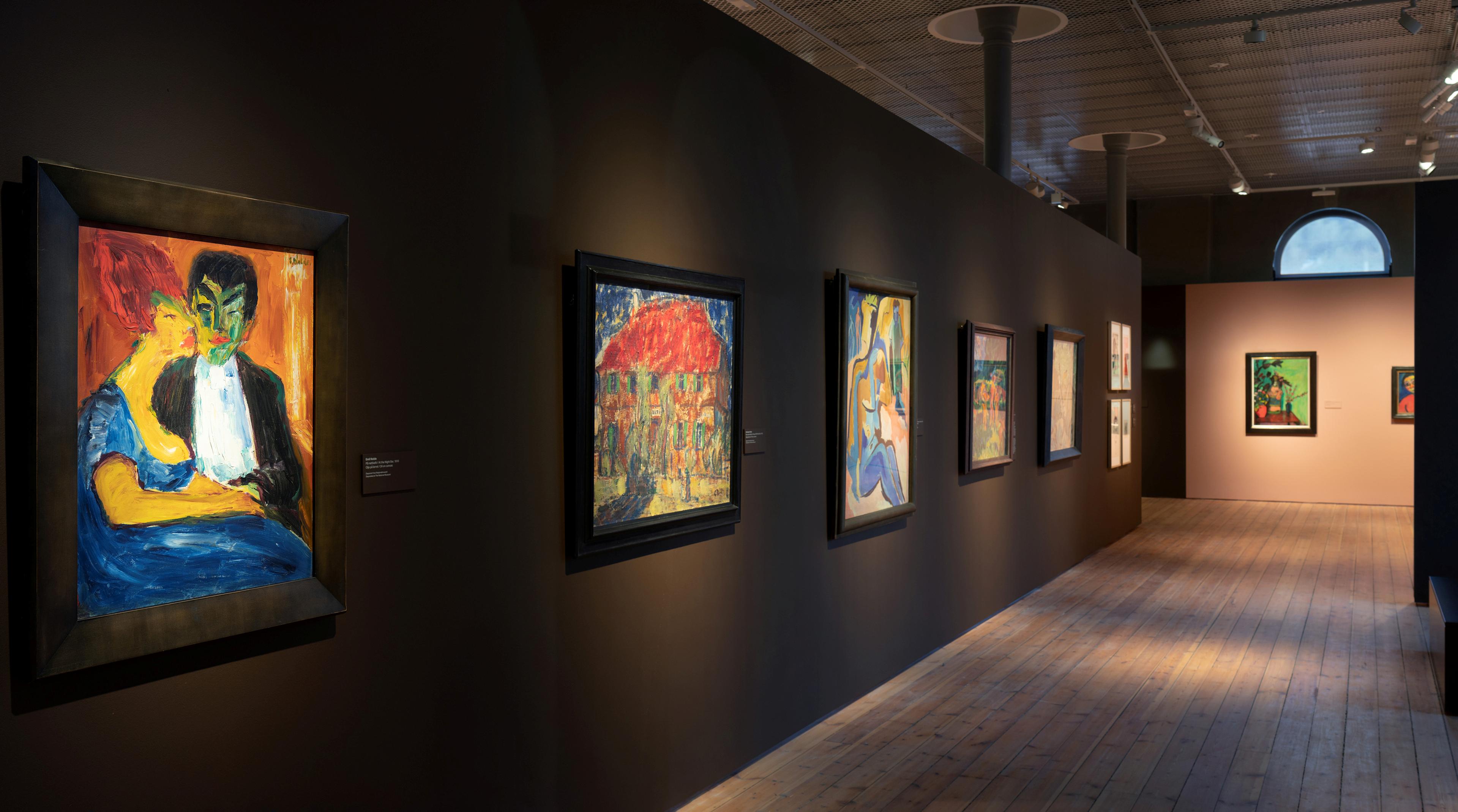 Fotografi fra utstillingslokalet som viser en rad med malerier på mørk vegg.