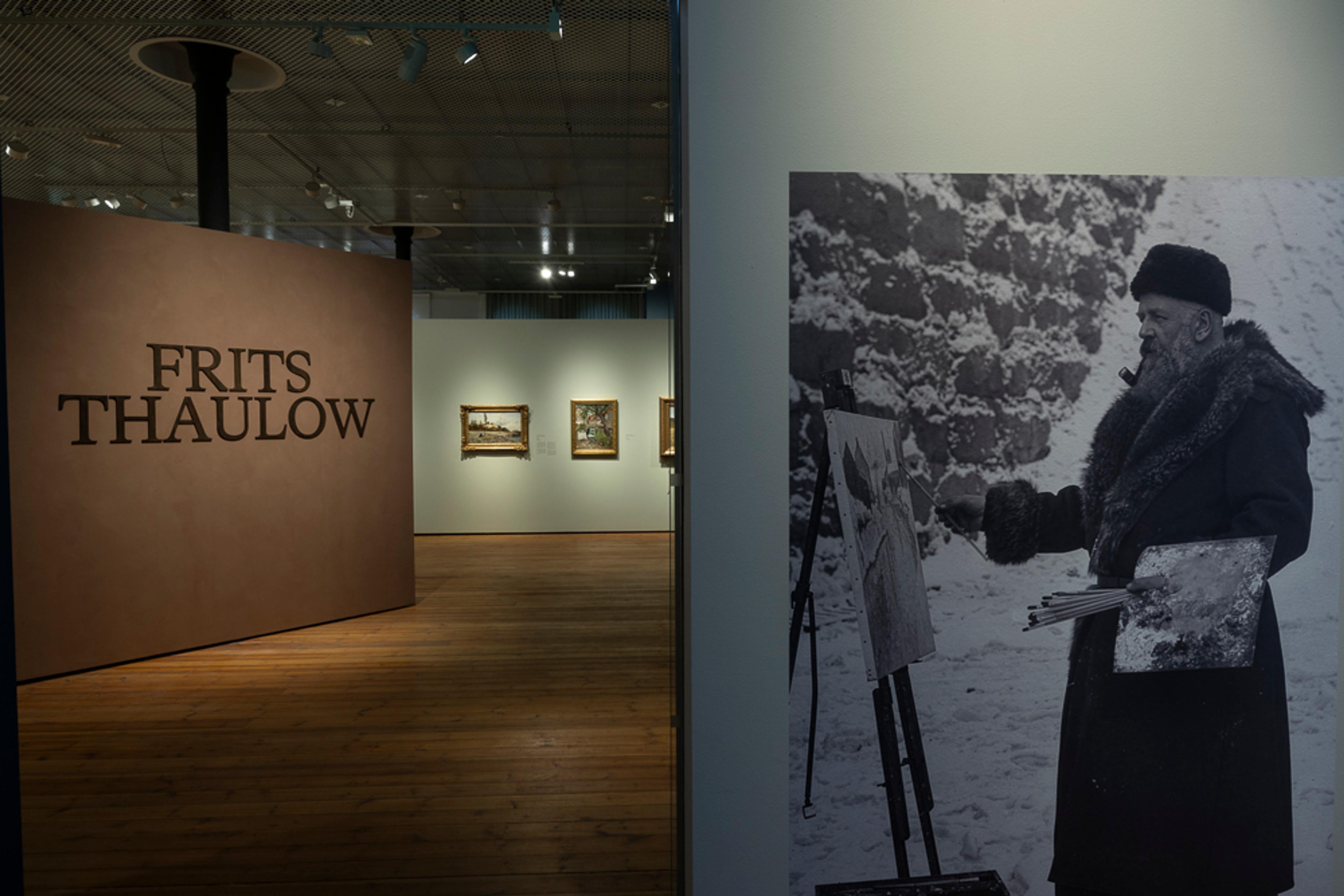 Et foto fra Thaulow utstillingen som viser malerier på vegg og en stor veggtapet med foto av kunstneren foran staffeliet