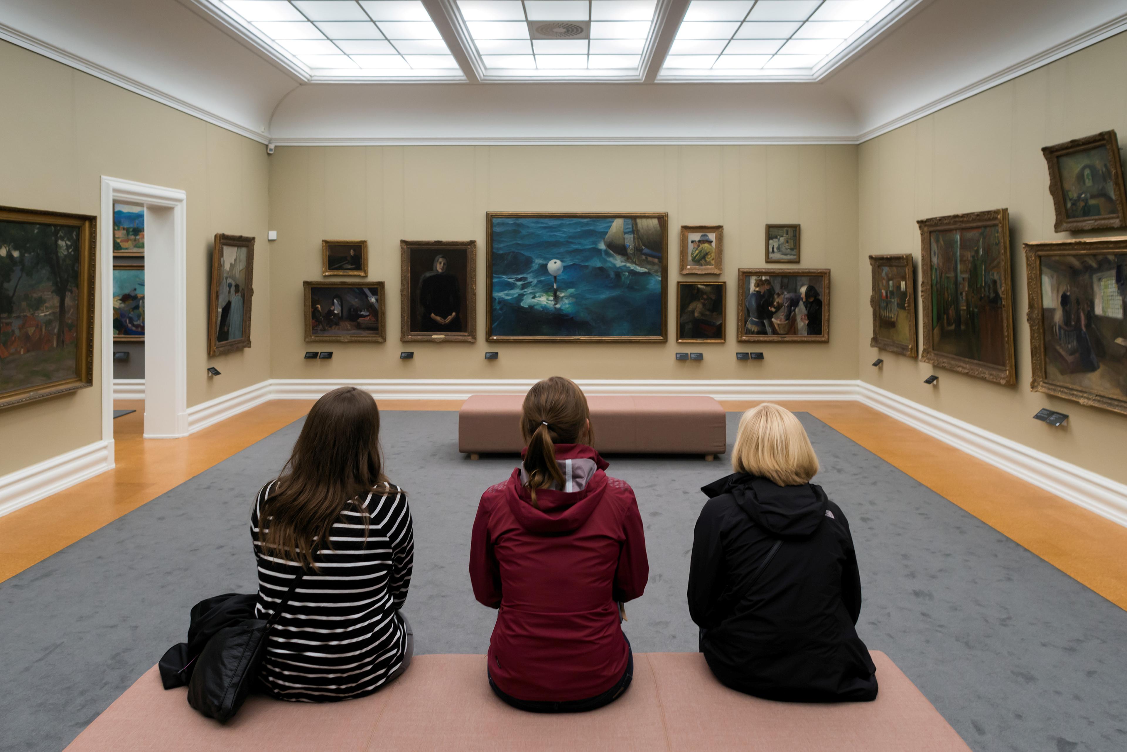 Three young people are sitting on a bench in the Rasmus Meyer collection, with paintings by Christian Krohg in the background.