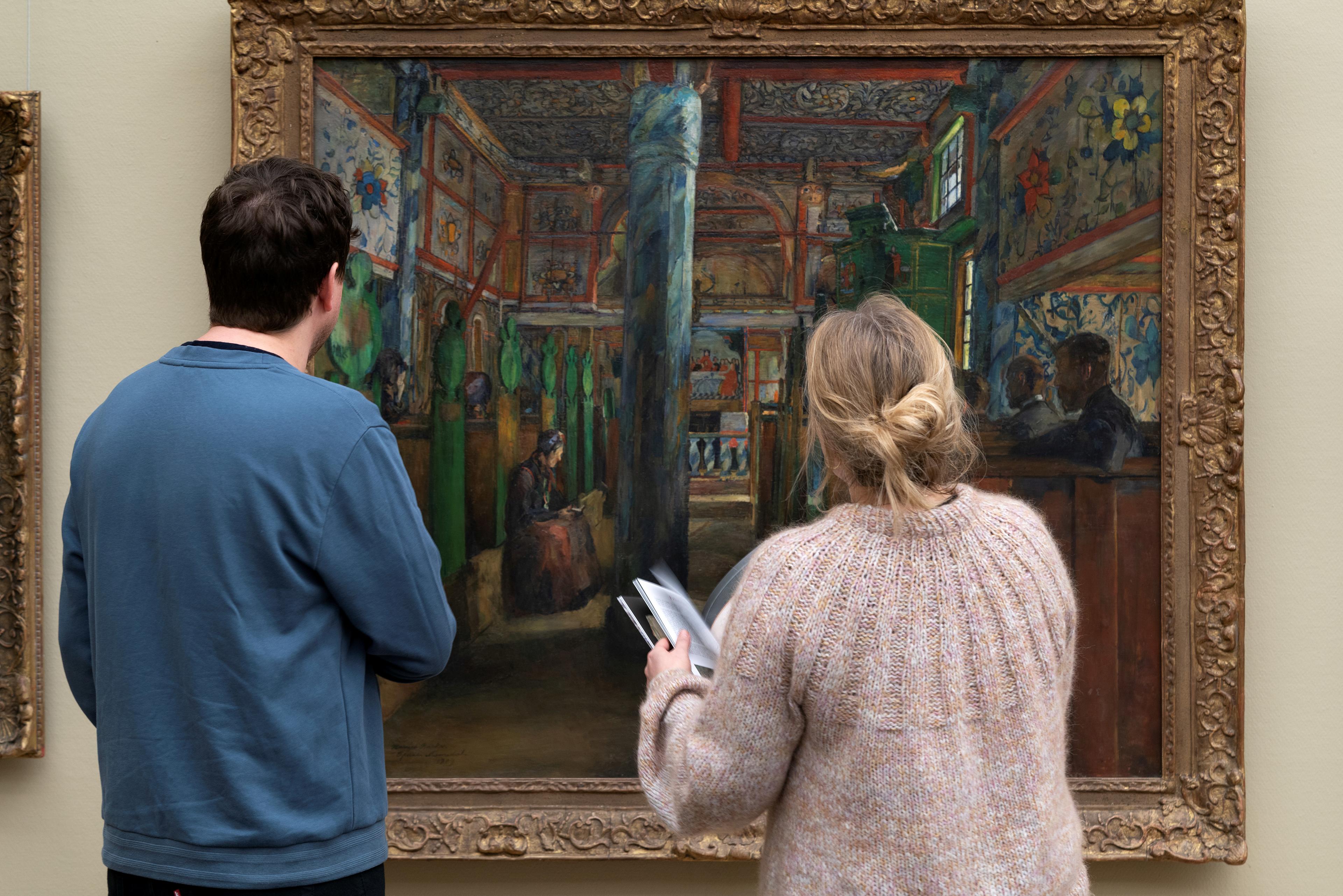 Two people are standing in front of a painting by Harriet Backer.