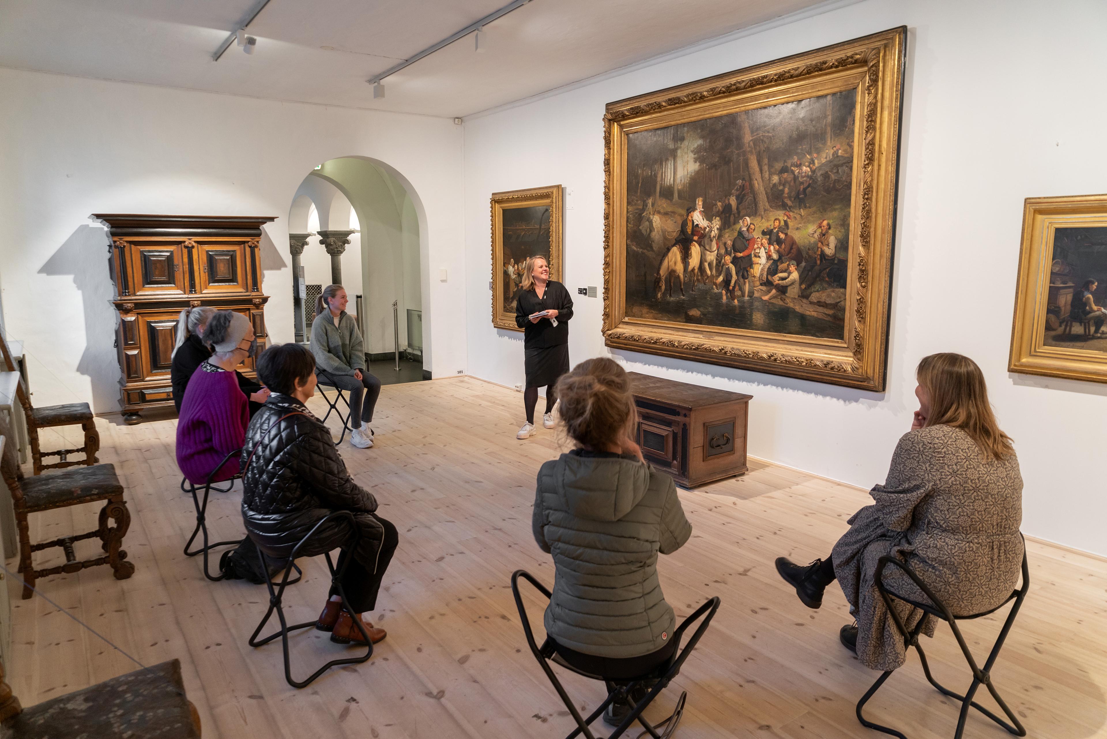 Foto av en formidler som snakker til en gruppe mennesker, mens de sitter foran et stort maleri i Rasmus Meyers samlinger.