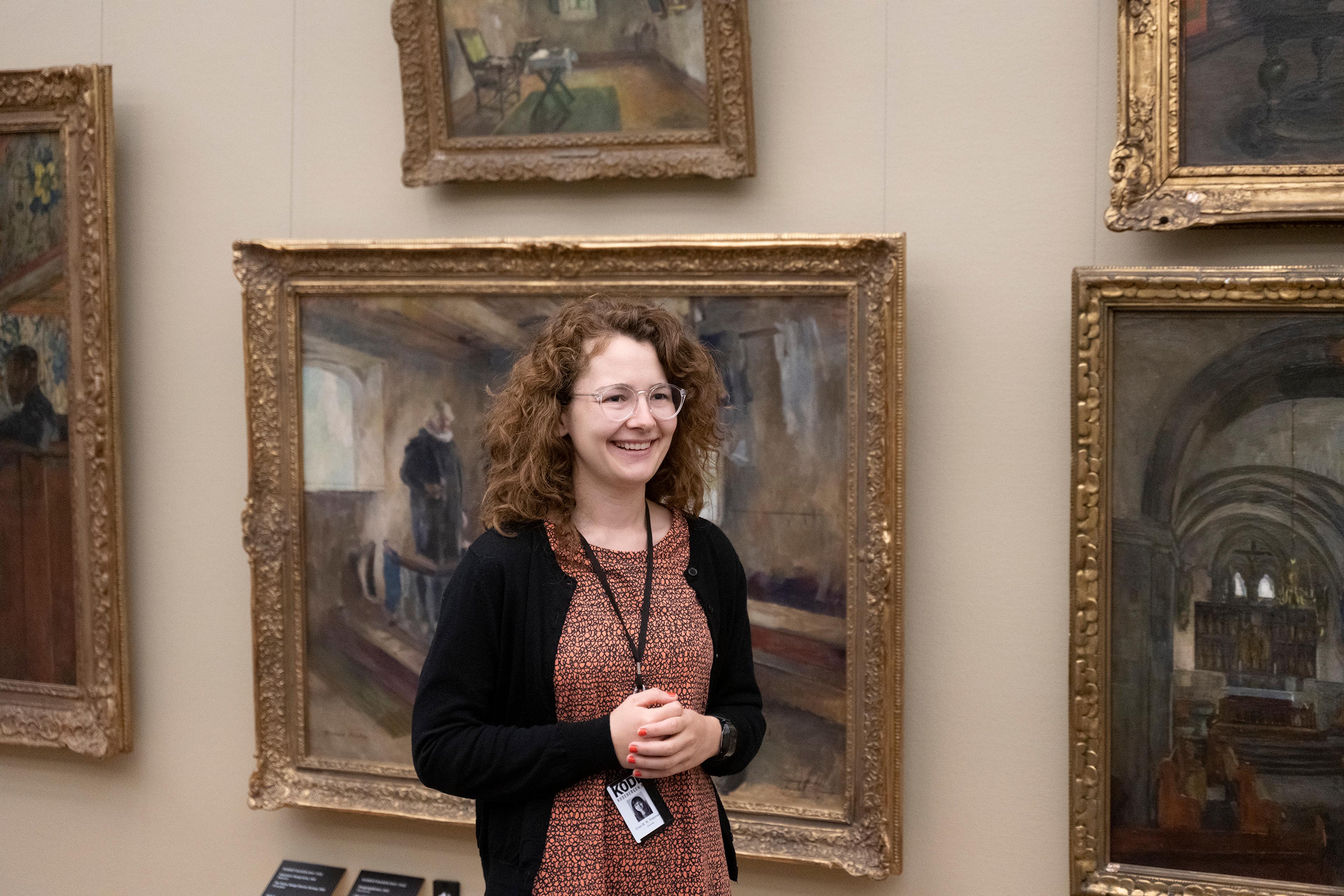 a woman is standing in front of a row of paintings, smiling