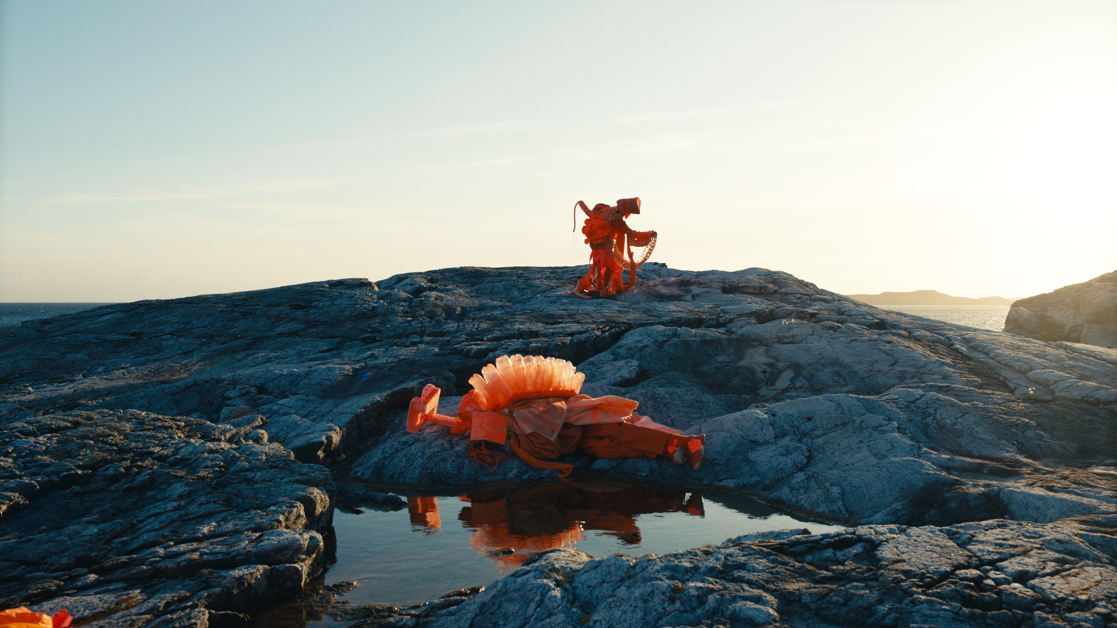 En videostill av dansende mennesker i oransje bølgende klær på en klippe