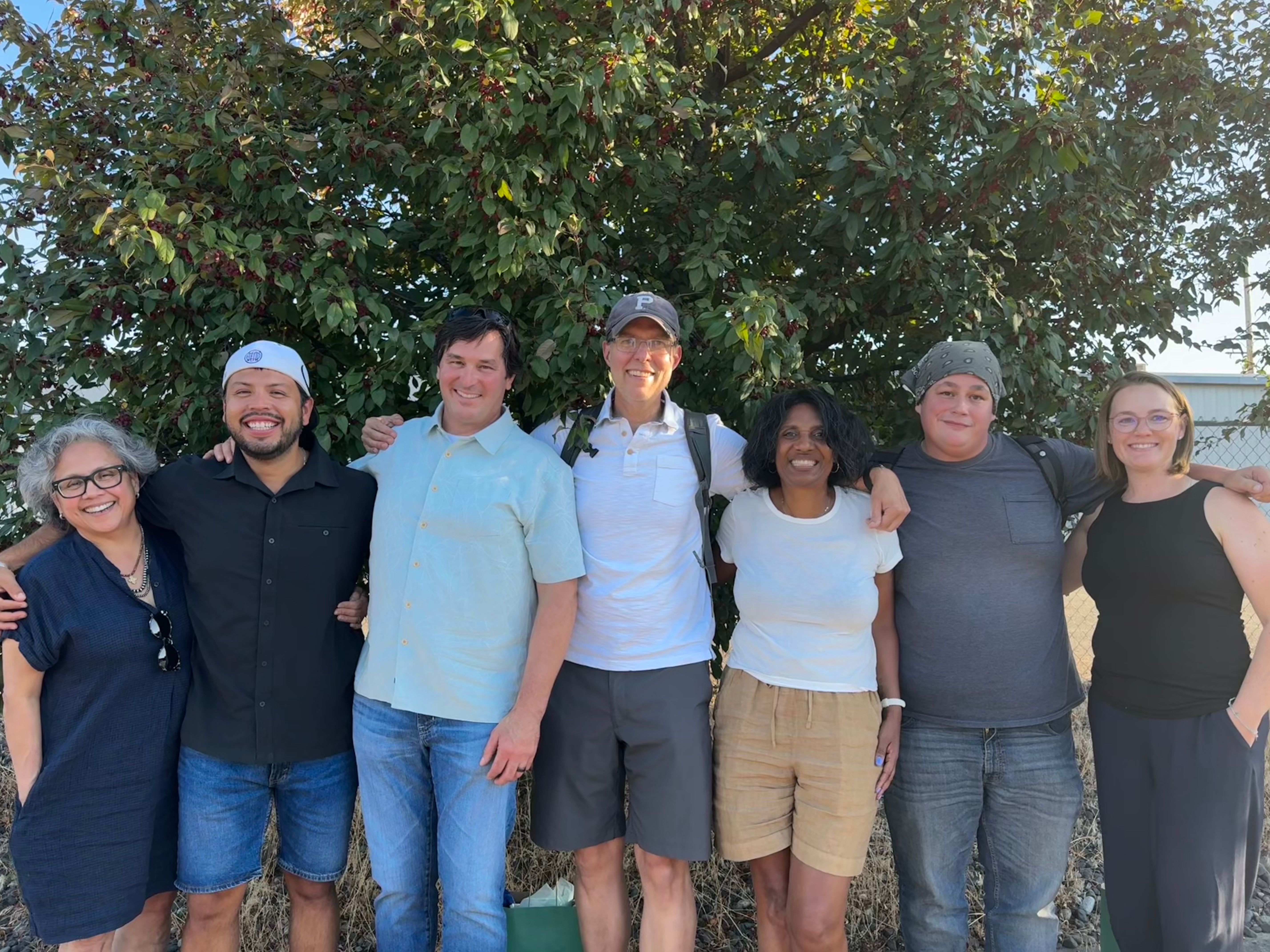 Seven members of our smiling board standing outside in front of a wall painted to resemble the edge of a forest.