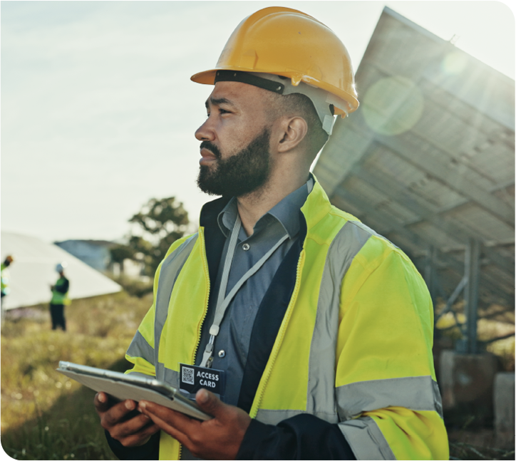 A field worker in a hard hat and safety vest holding a tablet beside a solar array.