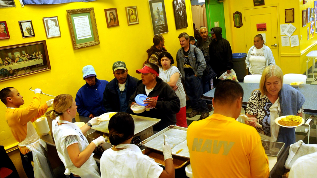 Volunteers serving hot meals to people queuing at a soup kitchen with bright yellow walls