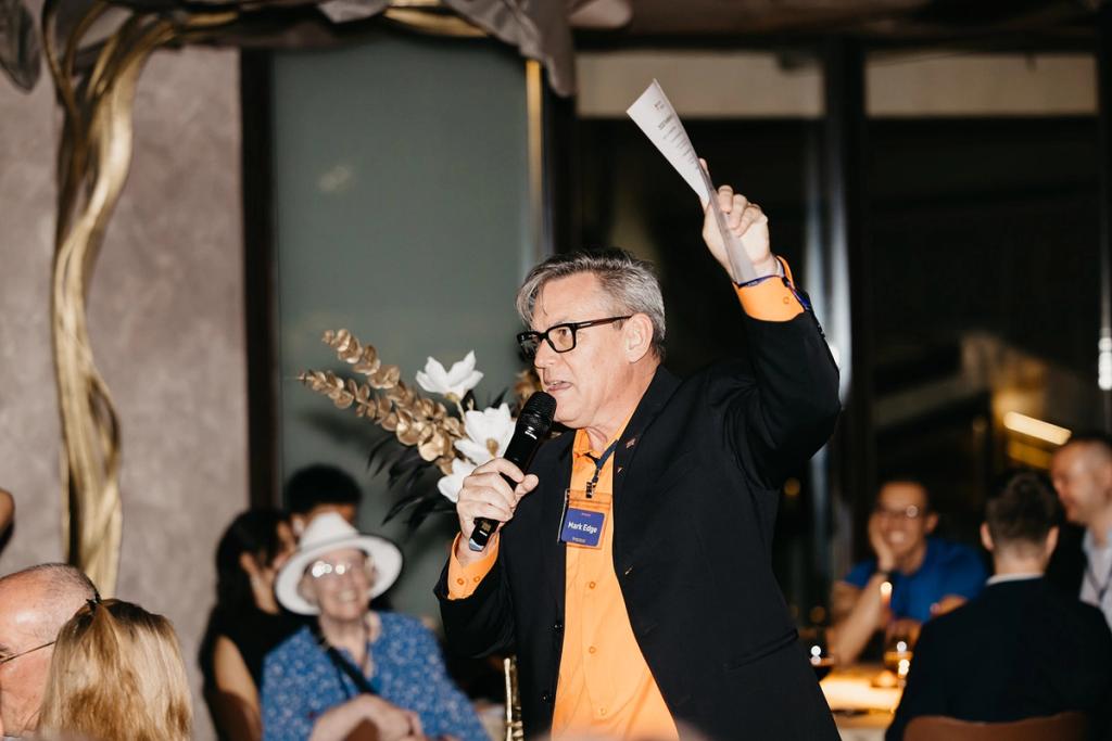 Mark Edge holding up a bid sheet with a microphone in hand among the tables of the VIP dinner