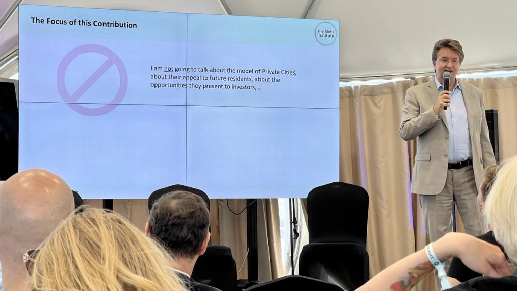 TIPOLIS CSO Dr. Andreas Baumgartner speaking at Zuzalu beside a slide on private cities