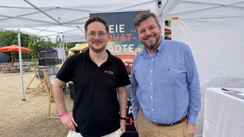 Two men at the Free Cities Foundation stand in front of a Freie Privatstädte banner