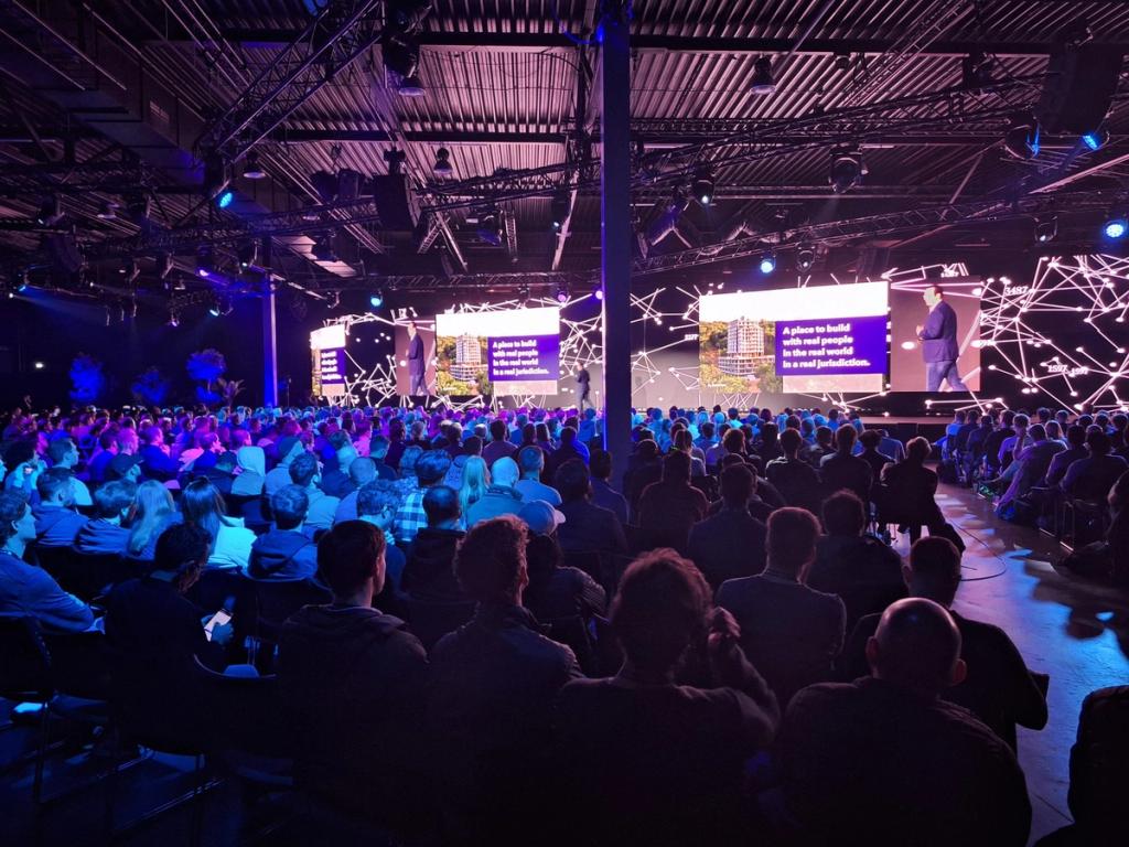 Full hall watching a Próspera talk with the slide A place to build with real people in a real jurisdiction
