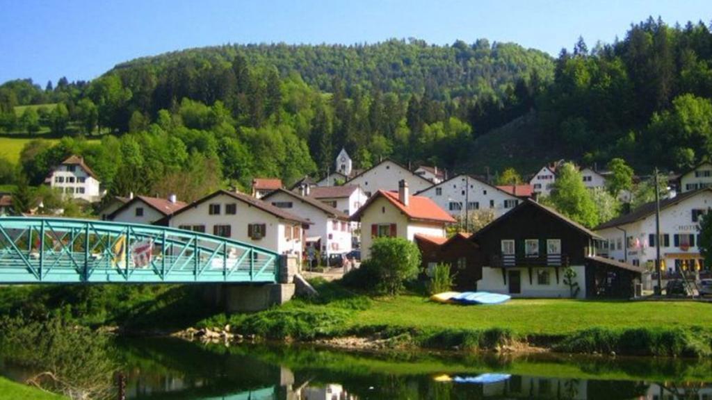 Swiss village of Soubey with white chalets, a green footbridge and a river below wooded hills
