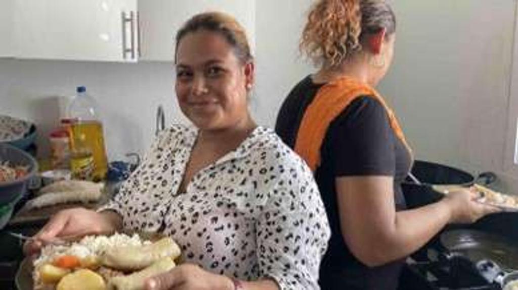 Rosa Aguilar holding a plate of food in her kitchen while an employee cooks at the stove