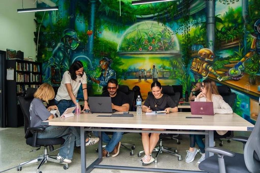 People working with laptops at a shared table in front of a colorful futuristic mural in a Próspera coworking space