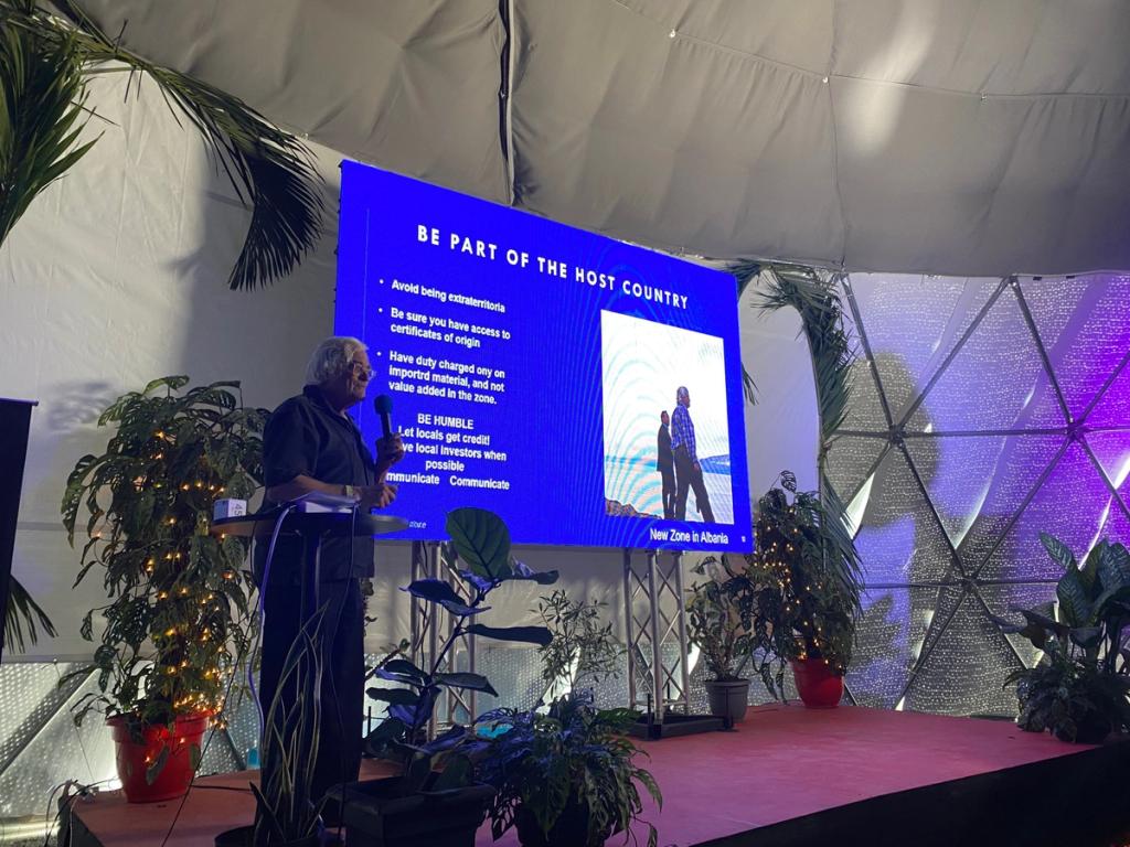Speaker in the Vitalia dome beside a slide titled Be part of the host country