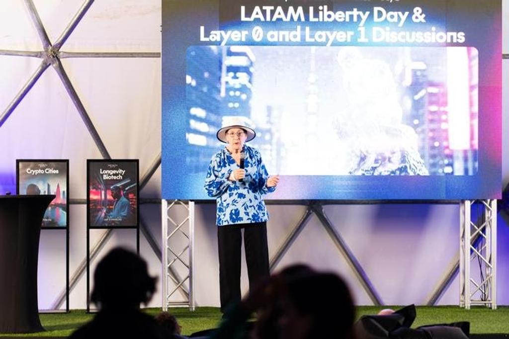 Speaker in a sun hat on stage under a LATAM Liberty Day screen at a summit in Próspera, Honduras