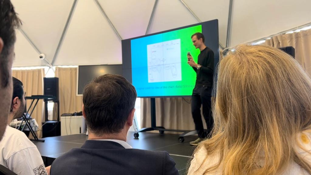 Vitalik Buterin presenting a chart on screen to the audience in a tent at the Zuzalu launch event