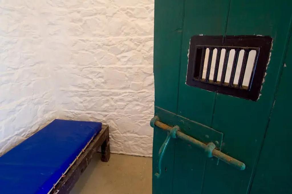 Spartan prison cell on Sark with whitewashed walls, a blue mattress bench and a green barred door