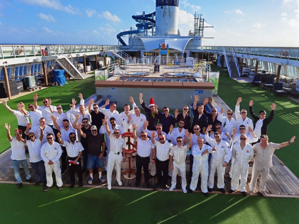 La tripulación del MS Satoshi, con uniformes blancos, saluda con los brazos en alto en la cubierta del crucero
