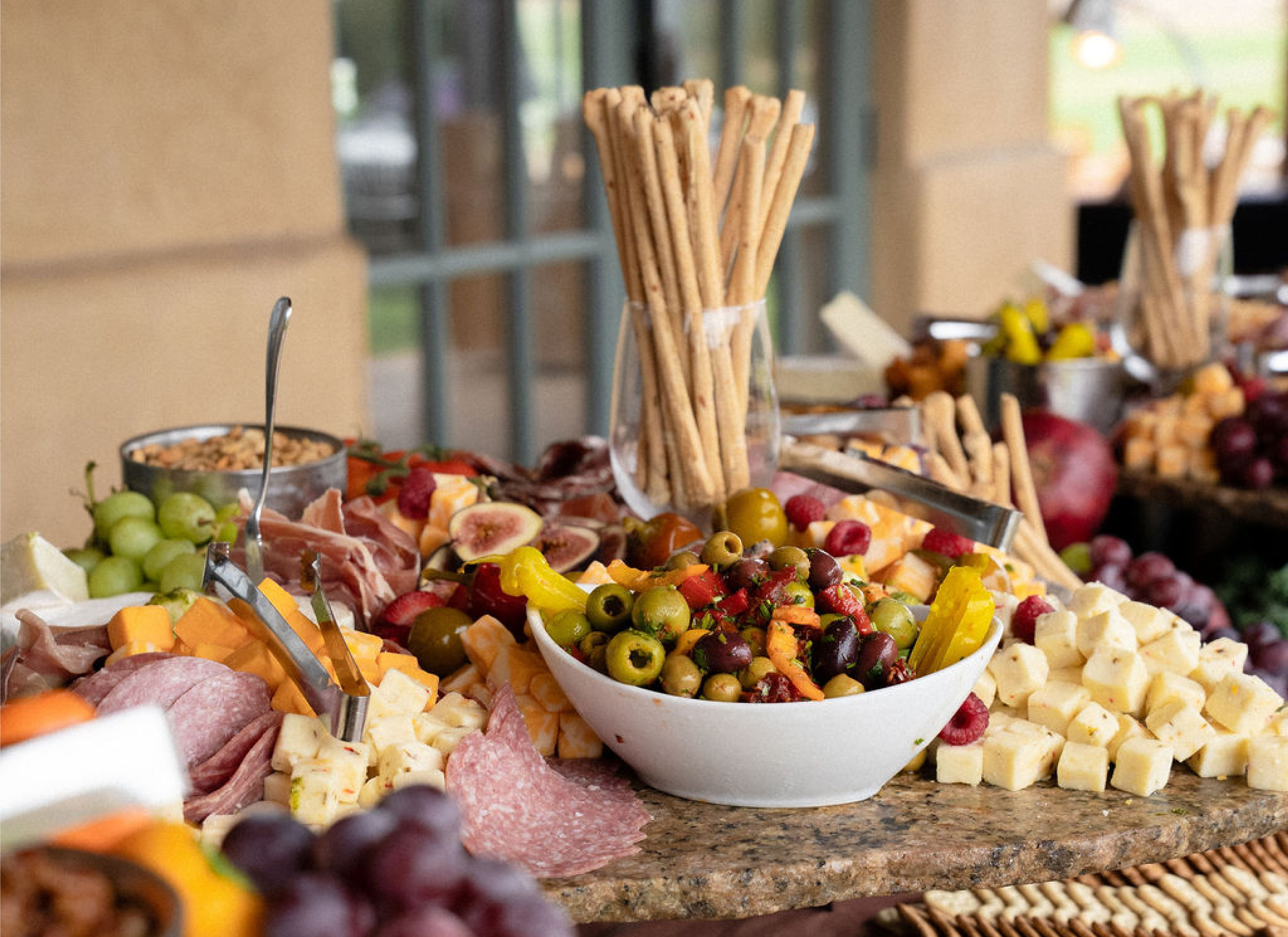 Colorful corporate catering grazing table with assorted cheeses, cured meats, olives, grapes, fruit, breadsticks, and appetizers arranged for an event.